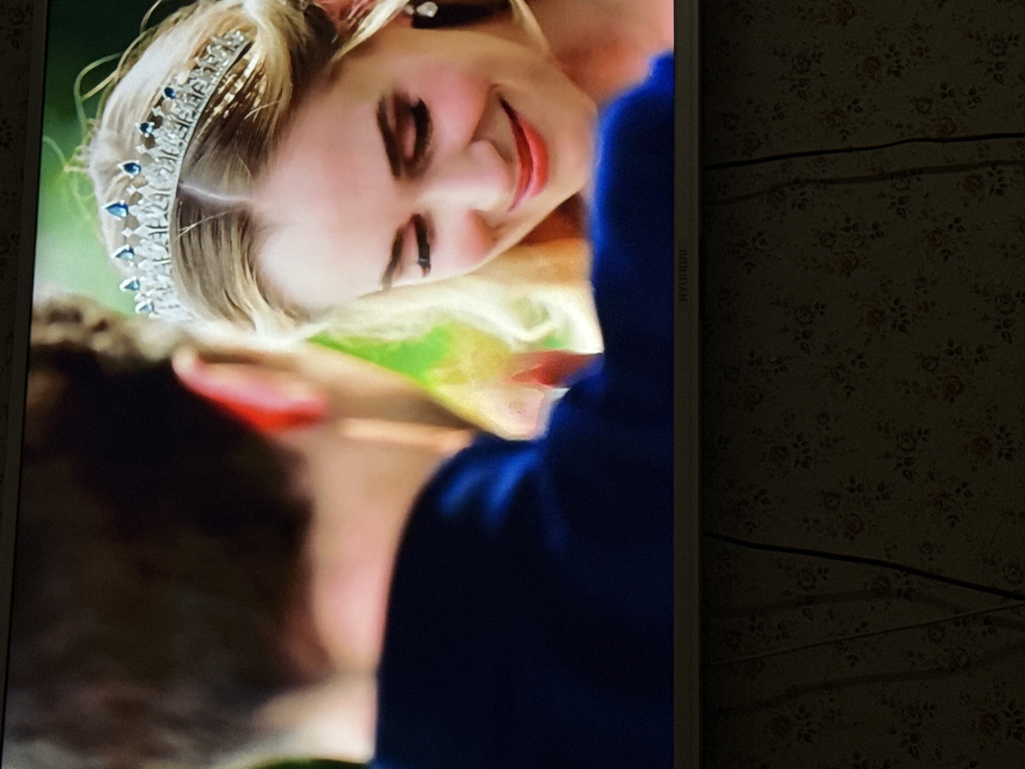 The image shows a television screen displaying a scene of a woman with blonde hair and a tiara, smiling and leaning towards a person with dark hair wearing a dark blue shirt. The woman has red lipstick. The television is a Hyundai brand. The background to the right of the television shows a wall with a floral pattern and two electrical cords. The image was taken indoors in a location unknown. There is no information to indicate it is in the city of Mytishchi, Russia.