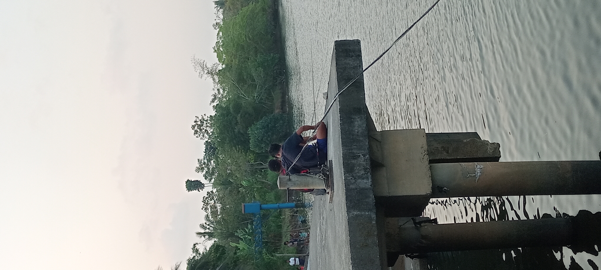 A wide shot captures a view from a concrete pier in Kalipucang, Indonesia, overlooking a calm body of water. The pier extends into the frame from the left, with two men sitting on the edge, facing away from the camera and towards the water. They are dressed casually, one in a dark t-shirt and shorts, the other in a black t-shirt and blue shorts. Both appear to be fishing, with fishing rods visible extending into the water.

In the background, a lush, green landscape of trees and vegetation lines the opposite bank of the water. The sky above is overcast and pale, suggesting either early morning or late afternoon. A blue water tower-like structure with a blue lid is visible on the left side of the pier, behind the two men. Further to the left, a group of three people, two adults and a child, are also visible on the pier, standing near the water's edge. The overall atmosphere is peaceful and tranquil, with the natural beauty of the surroundings contrasting with the man-made structure of the pier.