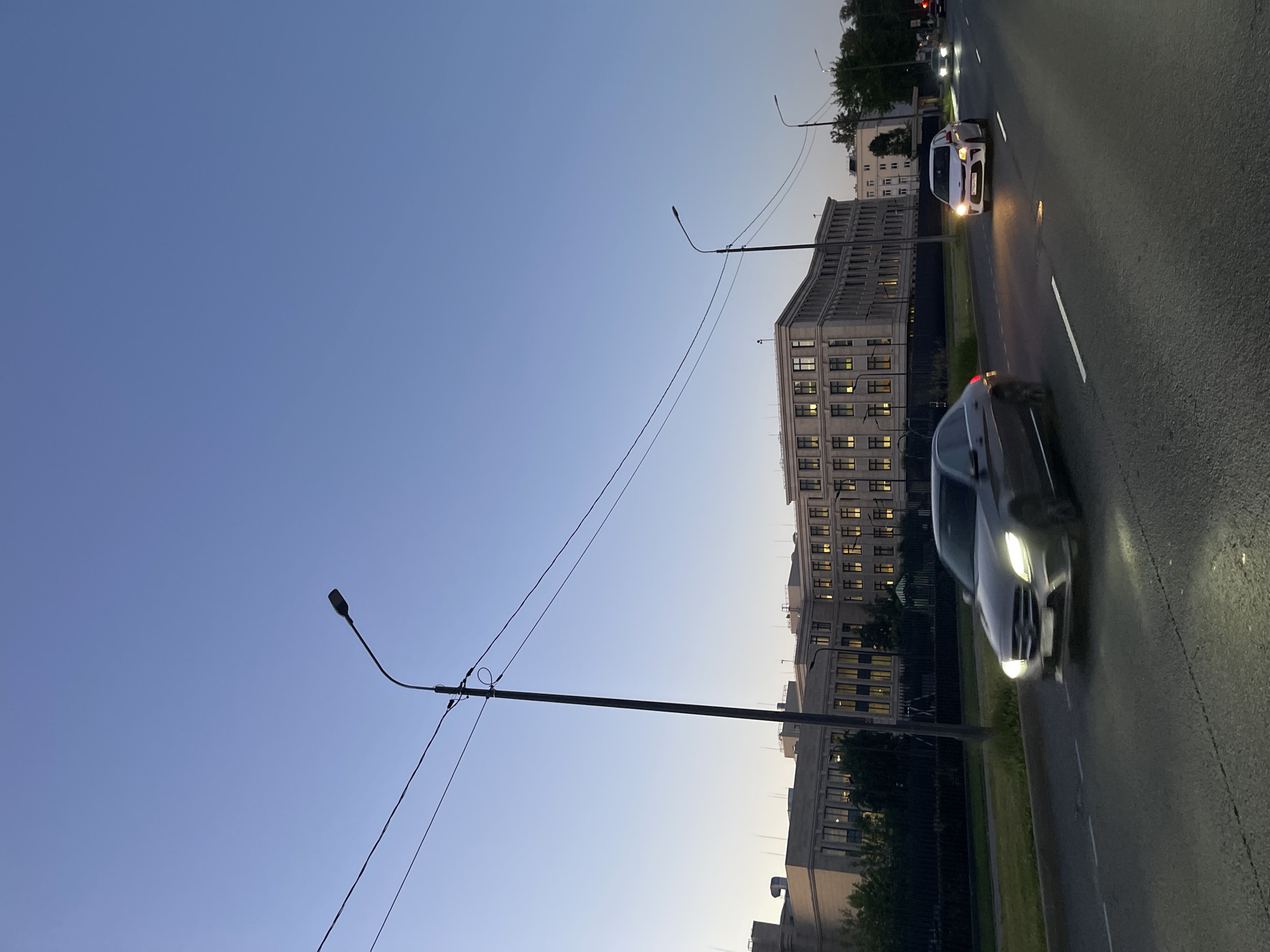 The image depicts a twilight scene on a city street, likely in Saint Petersburg, Russia, given the architectural style of the building. A wide asphalt road occupies the foreground, marked with white lane dividers. A gray sedan, possibly a Mercedes-Benz, is in motion on the left lane, its headlights illuminated. Further down the road, a white hatchback, possibly a Kia, is also visible, its headlights on. Several other vehicles are present in the distance.
To the left of the road, a paved sidewalk runs alongside a grassy area and a dark metal fence. Behind the fence, a large, multi-story building with a light-colored facade and numerous windows is prominent. The windows are illuminated, suggesting the building is occupied. Several streetlights are visible along the road, some with overhead wires. Trees and other buildings can be seen in the background. The sky is a clear, pale blue, transitioning to a darker shade of blue towards the top of the frame, indicating dusk or early evening. The overall atmosphere is one of urban activity during the transition from day to night.
