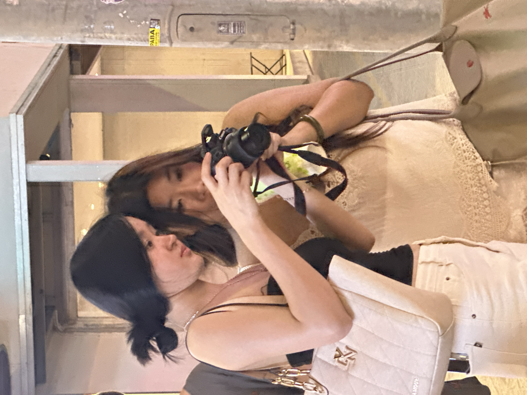 Two young Asian women are standing on a sidewalk in front of a building with a streetlight pole to the right. The woman on the left, with black hair pulled back in a ponytail, is wearing a black tank top and white pants. She has a quilted cream-colored handbag with gold chain straps and a gold "LV" logo. The woman on the right, also with dark hair, is wearing a light-colored lace top. She is holding a black digital camera and appears to be taking a photo. She is also carrying a light-colored handbag with a red logo. The environment suggests a commercial or entertainment district, possibly in Da Nang, Vietnam, given the presence of a "PAPA" sticker on the streetlight. The lighting is artificial, indicating it is either evening or night.