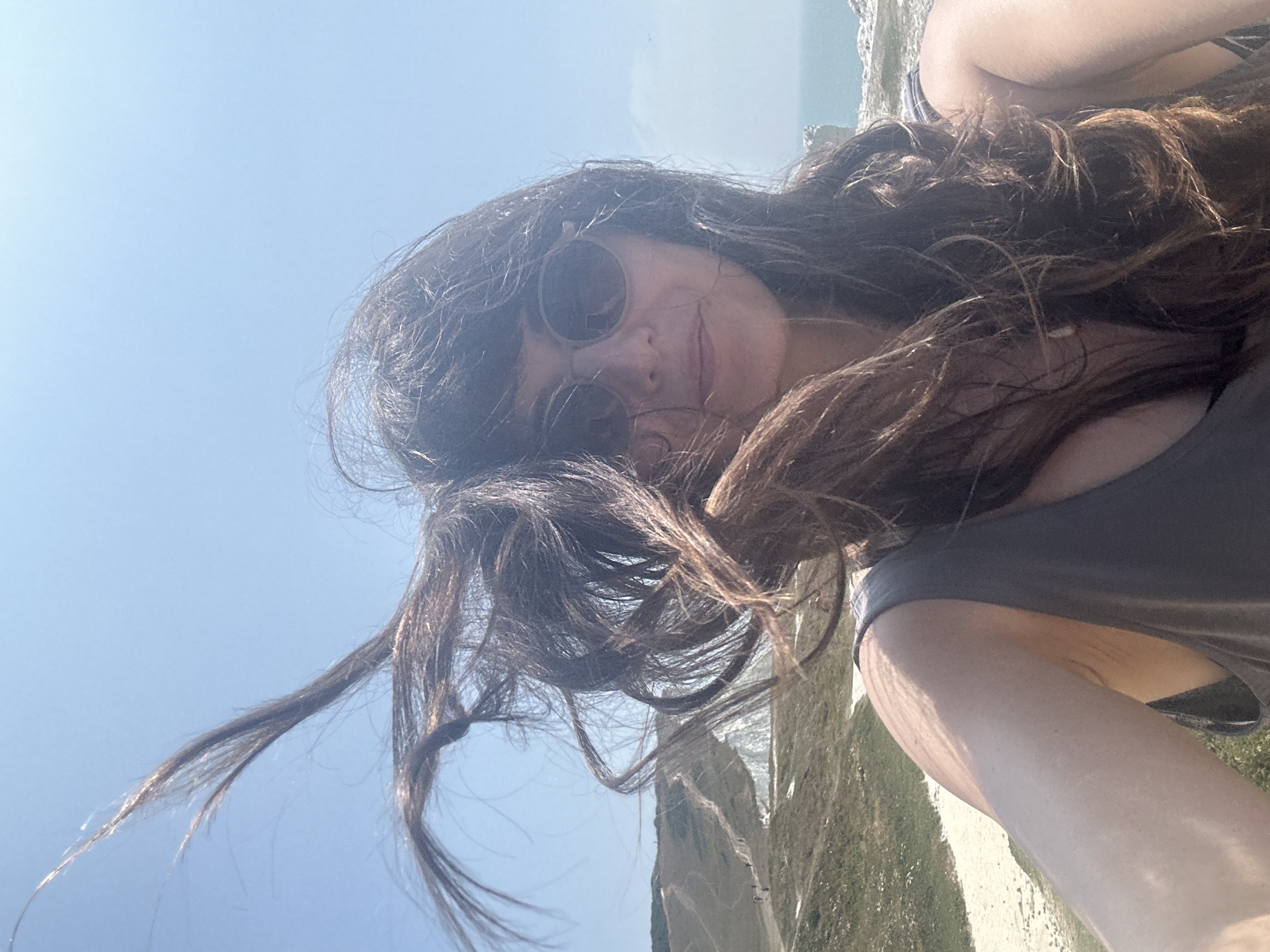A selfie is taken by a woman with long, dark, wavy hair blowing in the wind. She is wearing sunglasses and a dark grey tank top. She is standing outdoors with a bright blue sky behind her. In the background, there is a scenic view of rolling green hills with a path winding through them, and the sea in the distance. The image appears to be taken on the South Downs Way near Seaford, East Sussex, United Kingdom.
