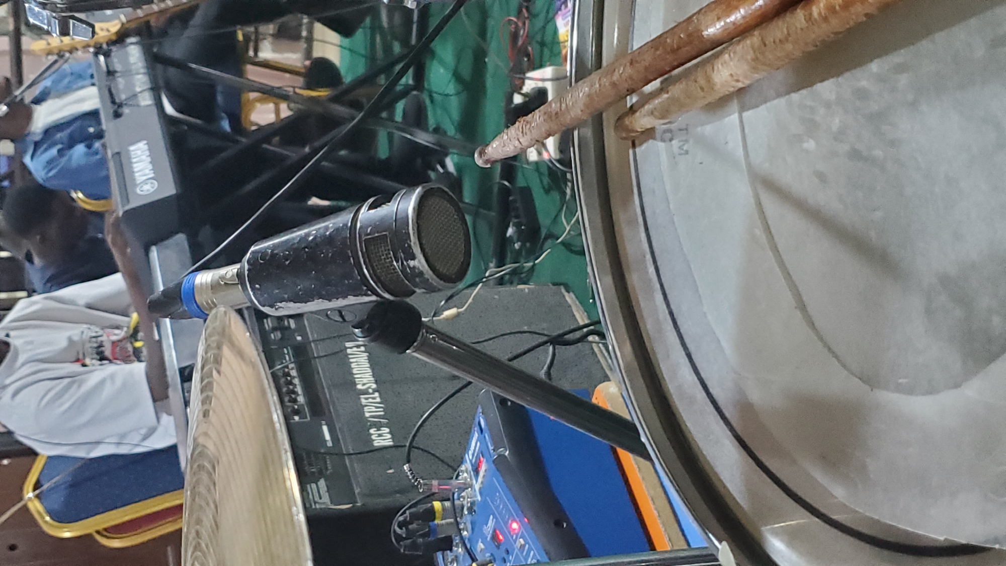 The image depicts a close-up view of musical equipment in what appears to be an indoor setting, possibly a church or performance space in Jalingo, Nigeria. In the foreground, a drumhead is visible with two drumsticks resting on it, one slightly overlapping the other. Behind the drumhead, a microphone on a stand is positioned prominently. To the left of the microphone, a blue audio mixer with several red indicator lights is present. In the background, a Yamaha keyboard is visible, along with people seated behind the instruments. One person is wearing a white t-shirt with a graphic design. The overall impression is of a band preparing for or in the midst of a musical performance.