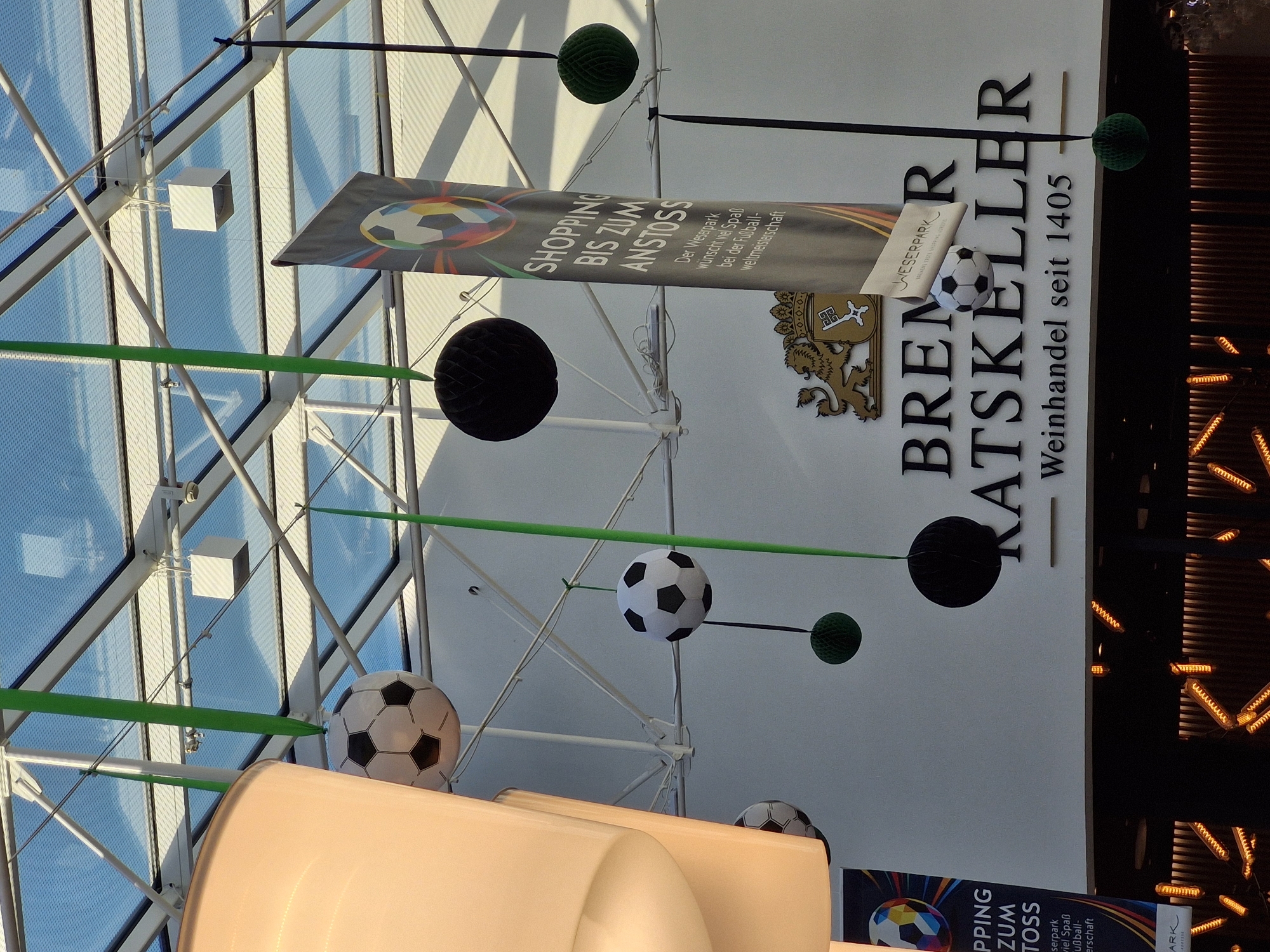 The image shows a close-up view of a shopping center interior, likely in Bremen, Germany, with a ceiling made of glass panels supported by a white metal structure. Several soccer balls, both real and decorative, are suspended from the ceiling by green and black ribbons, along with black and green honeycomb balls. A large banner with the text "SHOPPING BIS ZUM ANSTOSS" and a soccer ball graphic hangs prominently. Below the banner, the words "BREMER RATSKELLER Weinhandel seit 1405" are visible on a white wall, indicating the establishment. The lighting appears to be a mix of natural light filtering through the glass ceiling and artificial lights from below, some of which have a distinctive coiled filament design. There are no people visible in the frame. The overall atmosphere suggests a festive or event-based decoration, possibly related to a football (soccer) championship.