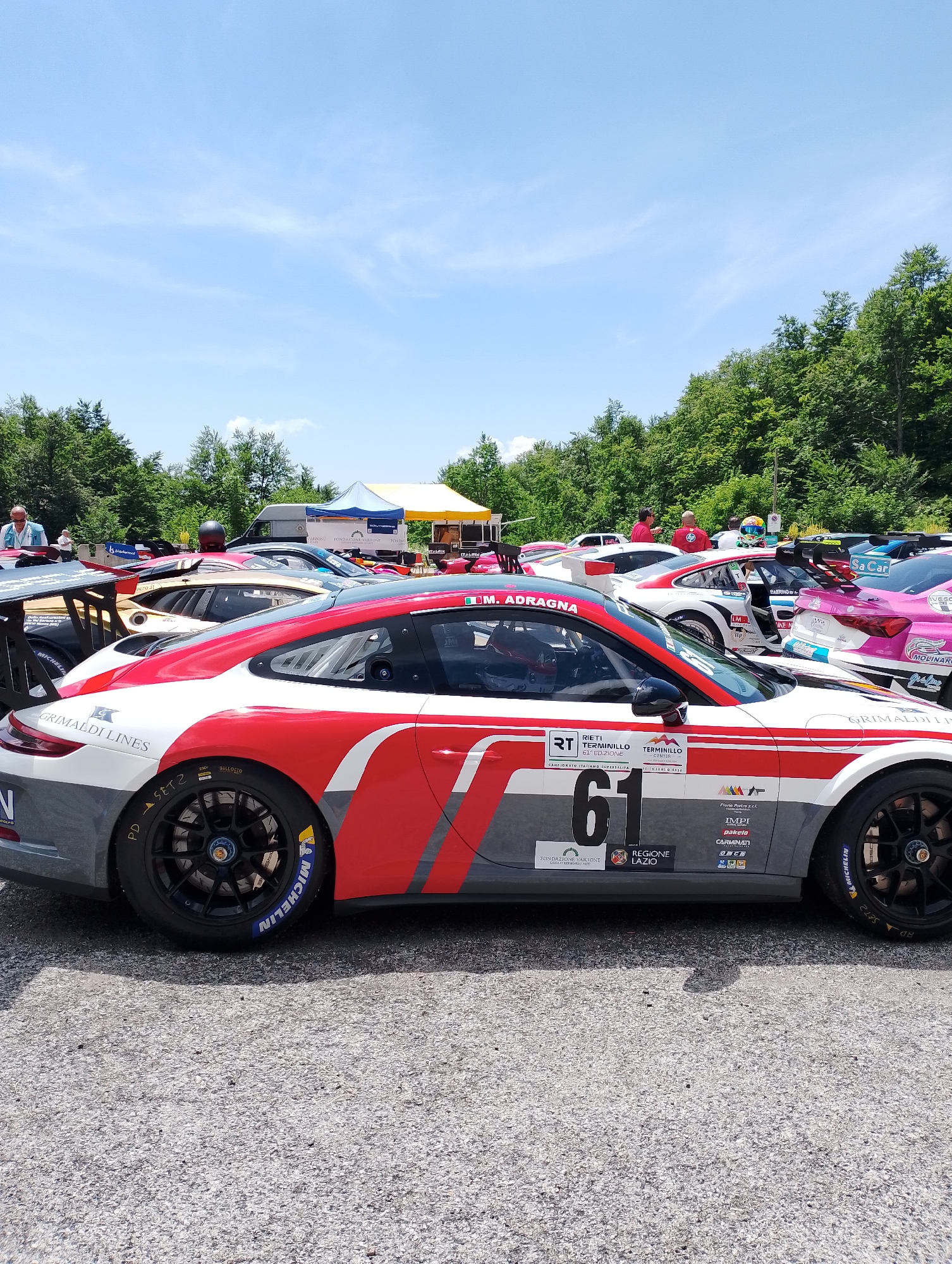 The image captures a scene at the Rieti racetrack in Italy, specifically during what appears to be a racing event. In the foreground, a red and white Porsche race car with the number "61" prominently displayed is parked on a gravel surface. The car features various sponsor logos and graphics, including "GRIMALDI LINES" and "REGIONE LAZIO." Its tires are labeled "MICHELIN." In the background, other race cars are visible, including a pink car and a gold and black car. Several people are standing around the cars, some dressed in casual attire and others in racing gear. Tents and temporary structures are set up behind the vehicles, suggesting a pit or paddock area. The environment is outdoors, with a clear blue sky overhead and a dense line of trees forming a backdrop. The overall impression is of a busy, organized motorsport event.