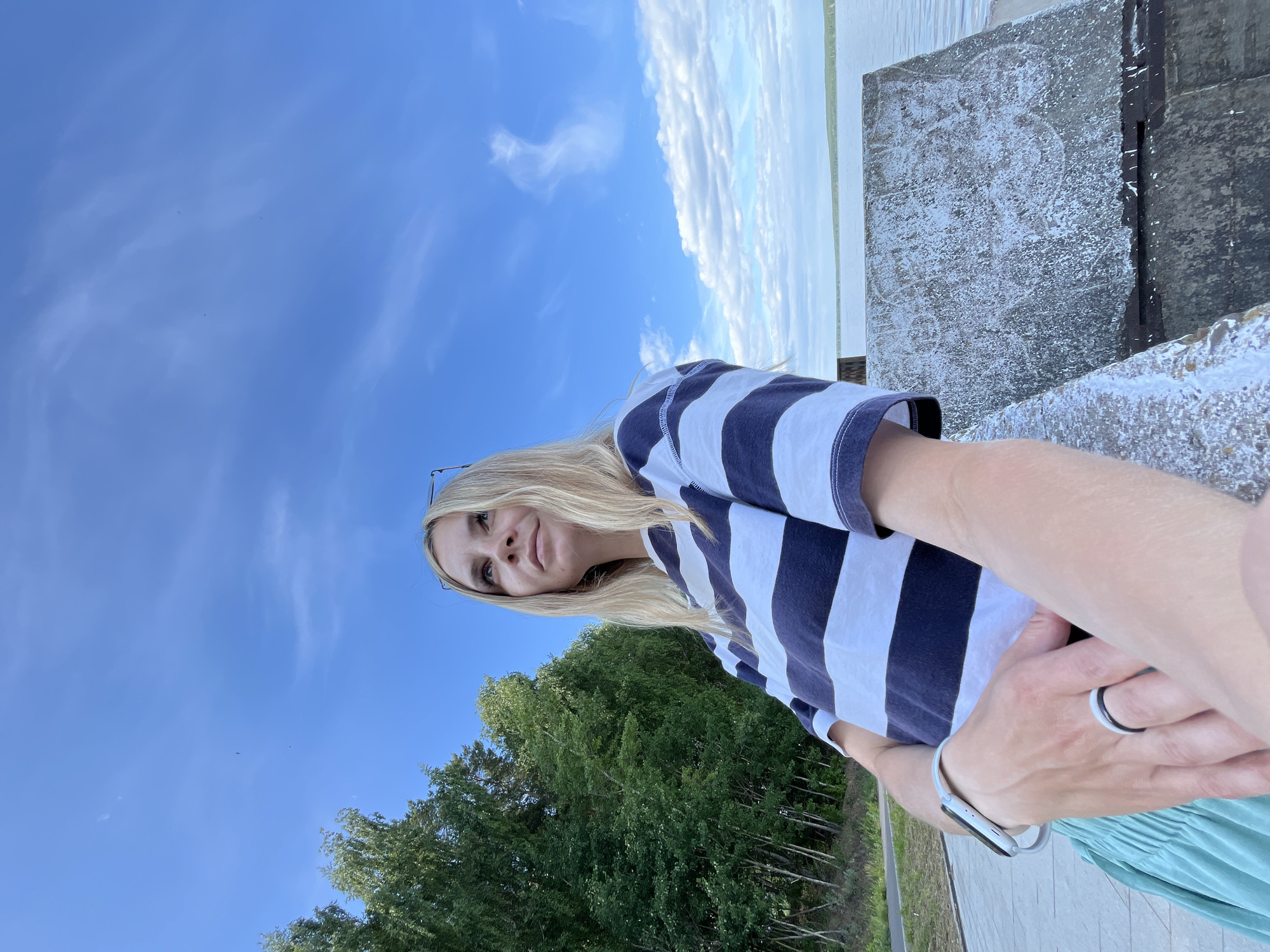 An outdoor, low-angle portrait features a fair-skinned individual with long blonde hair, wearing a navy blue and white striped t-shirt, a light green pleated skirt, and sunglasses perched on their head. The individual is looking upward and to the right, with a thoughtful expression. A white smartwatch is visible on their left wrist, and two rings (one black, one white) are on the left ring finger. The left arm rests on a weathered concrete structure, which is partially obscured and shows white flaking material and faint circular markings. The background consists of a clear blue sky with scattered cirrus and cumulus clouds. To the left, a dense stand of green deciduous trees is visible, while to the right, a large body of water extends to a distant treeline. Light-colored paving stones are visible on the ground in the lower left. The scene is located in Chaikovsky, Russia.