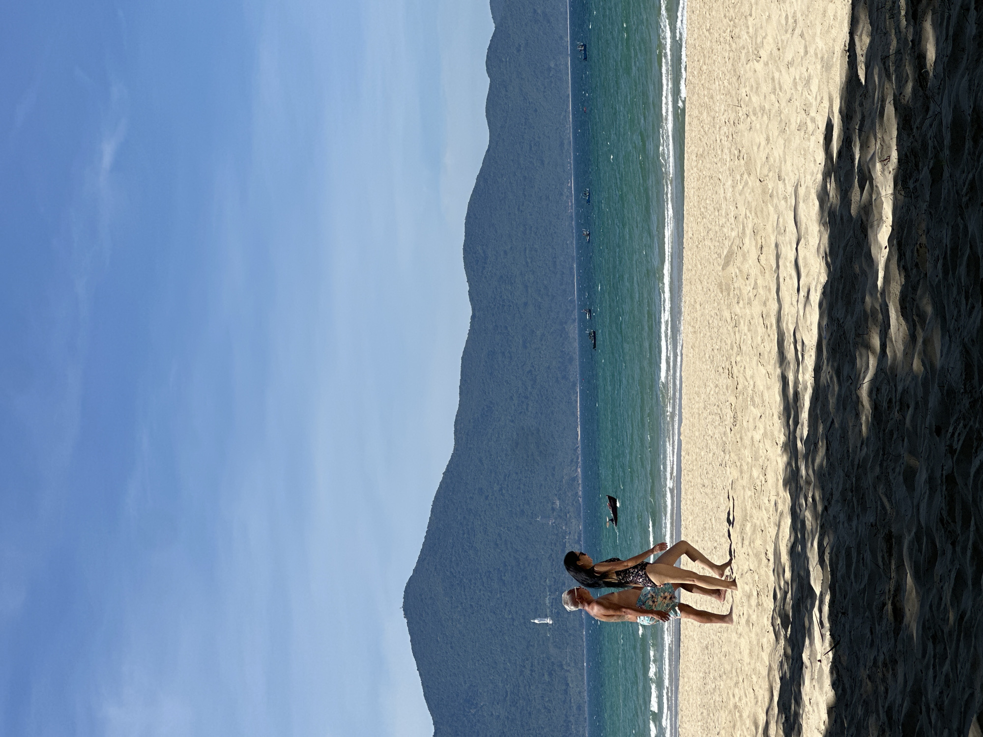 In Đà Nẵng, Vietnam, on a sunny day, a couple walks along the sandy shore of a beach. The man, with gray hair, wears floral patterned shorts, and the woman, with long dark hair, is in a patterned one-piece swimsuit. They are walking to the right, away from the viewer, side-by-side, with the sea to their left and the sand to their right. The ocean is a vibrant teal, with moderate waves breaking and several small boats scattered on the horizon. A large, tree-covered mountain range dominates the background, with a white structure, possibly a statue or monument, visible on its slopes. The foreground of the beach is sandy, with dappled shadows from unseen trees overhead cast across the sand. The sky is a clear, bright blue with a few wispy clouds.