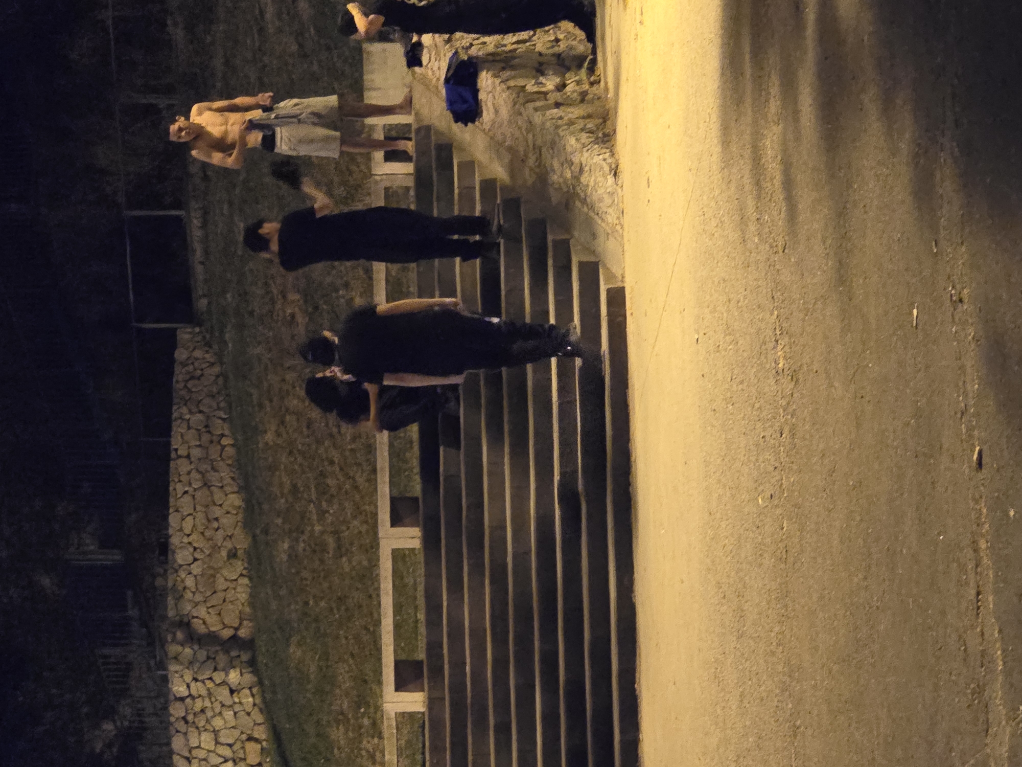 A group of young men are gathered on a set of concrete stairs at night. The man at the top of the stairs is shirtless and wearing shorts. Another man is standing next to him, holding a shirt. Two other men are on the stairs, wearing black shirts and pants. The environment appears to be an outdoor urban setting with a stone wall and grassy embankment behind the stairs. The scene is lit by artificial lights. It is implied that this scene is taking place in Cornellà de Llobregat, Spain.