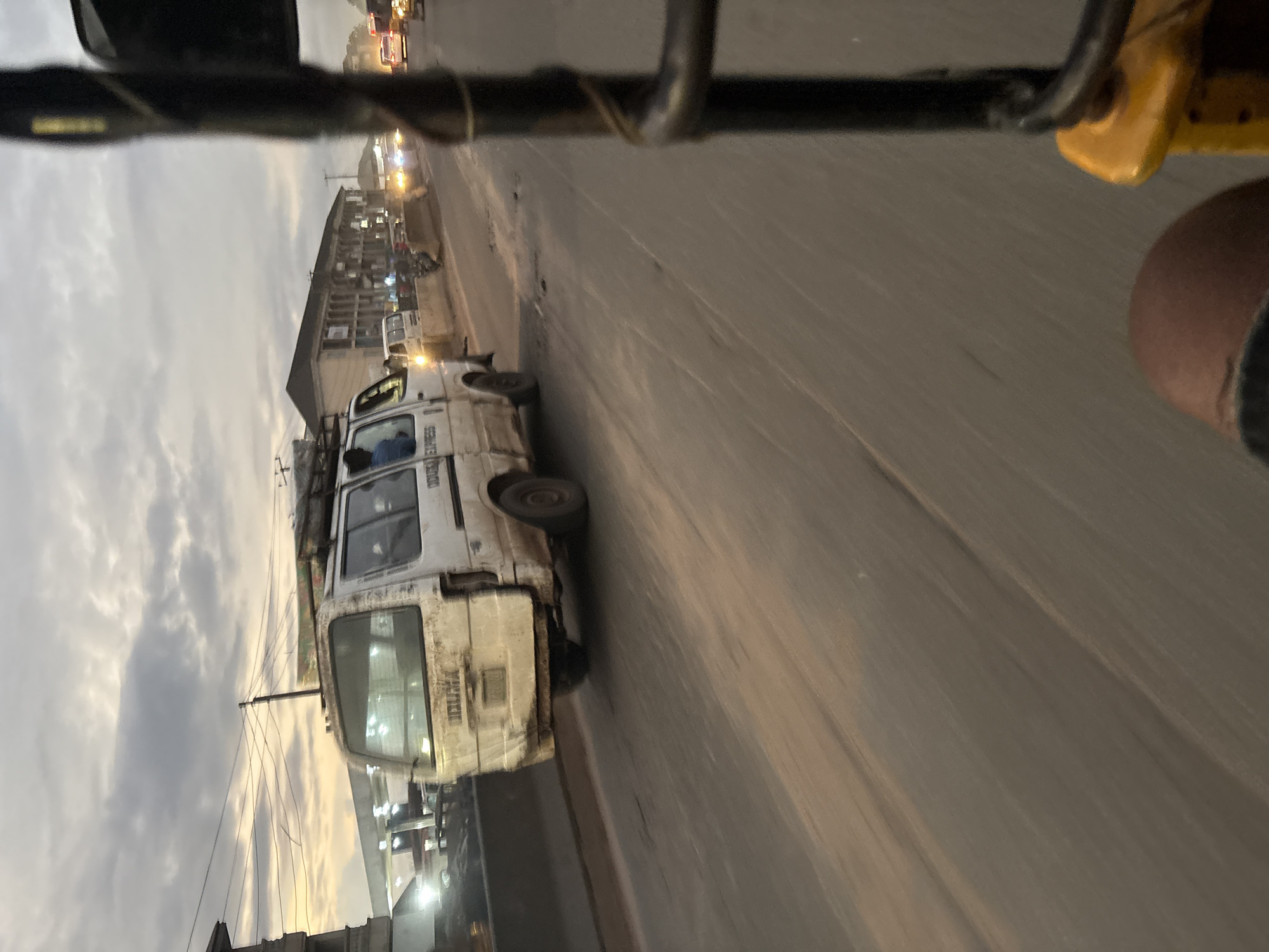 The image captures a street scene in Onitsha, Nigeria, during what appears to be dusk. The foreground features the leg of an individual, likely the photographer, and a portion of a yellow vehicle, possibly a rickshaw or a similar mode of transport. In the middle ground, a white minibus with "UDOMA EXPRESS" written on its side is prominently visible. The minibus appears weathered and has luggage strapped to its roof. Several other vehicles, including cars and what might be another minibus, are present on the road, suggesting moderate traffic. The road surface is asphalt, with visible tire marks and some debris. Buildings line the street in the background, with streetlights illuminating parts of the scene. The sky is overcast with dramatic clouds, tinged with the warm hues of a setting sun. The overall impression is of a bustling urban environment in the late evening.
