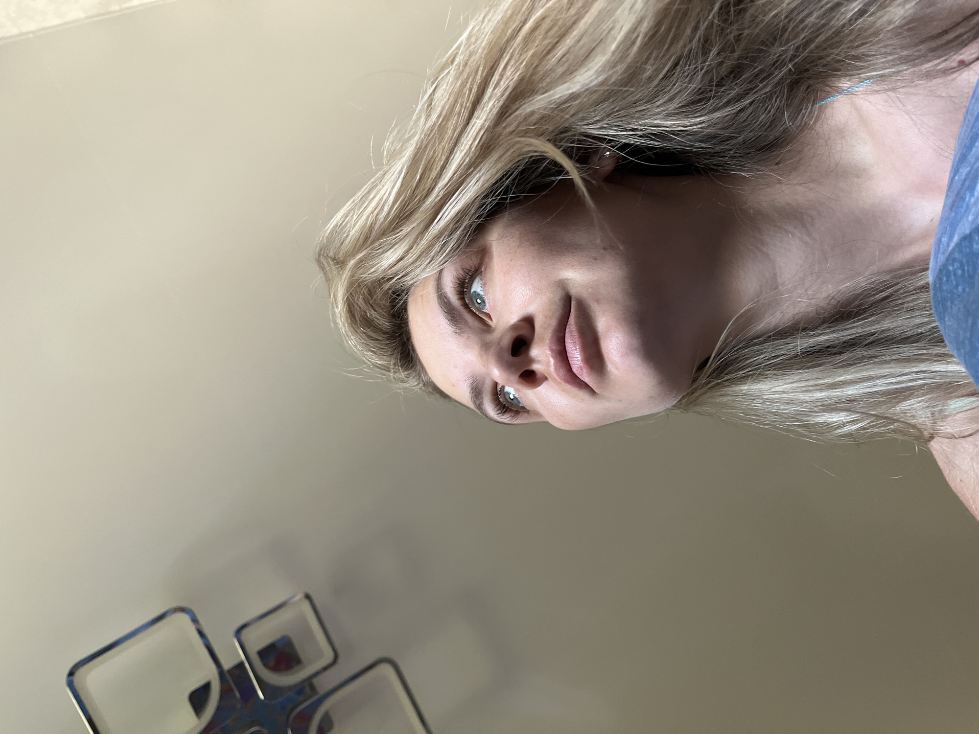 A low-angle, indoor close-up shot focuses on a fair-skinned woman with blonde hair and blue eyes. Her face is turned slightly towards the right. She wears a blue top. The background consists of a neutral-colored wall, and partially visible at the lower left corner are the frames of several squares. There is no information to identify the location as Chaikovsky, Russia.