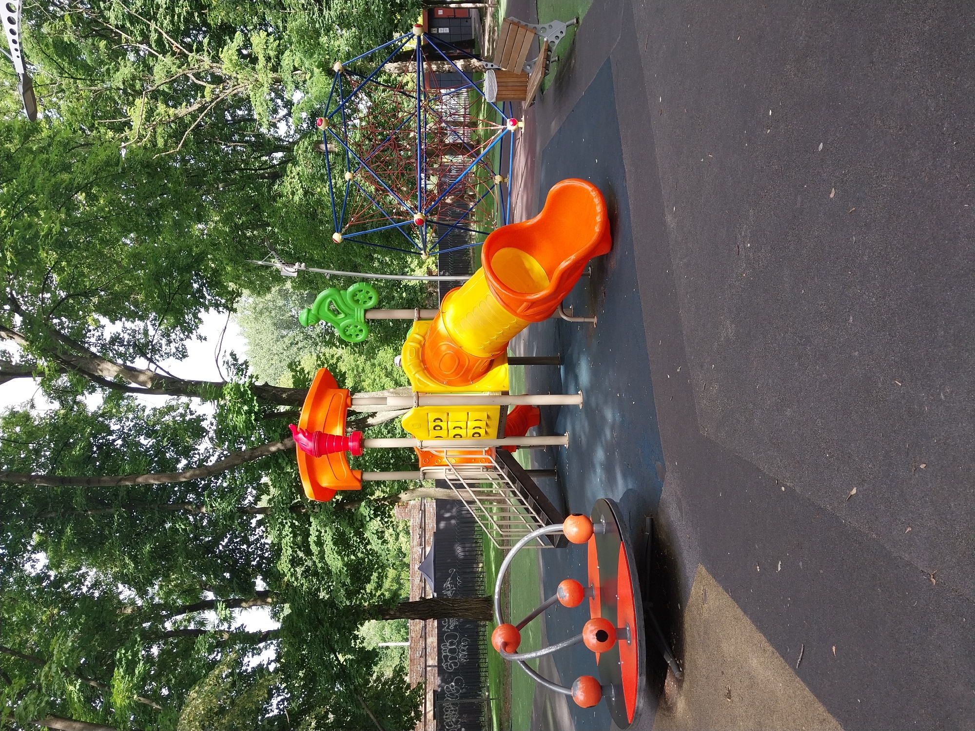 The image depicts a children's playground located in the city of Dnipro, Ukraine, likely in a park or residential area. The playground features a vibrant orange and yellow multi-level play structure with a slide, stairs, and various colorful panels. To the left, a spinning amusement ride with orange spheres is visible. In the background, a large geodesic dome climbing structure with blue netting and red accents stands out, adjacent to a wooden bench and a trash receptacle. The entire playground area is surfaced with dark rubber matting and some lighter-colored sections. Lush green trees surround the playground, suggesting a natural or park-like setting. The scene is captured from a slightly low angle, with the bright play equipment contrasting with the natural greenery and sky. There are no people visible in the image.