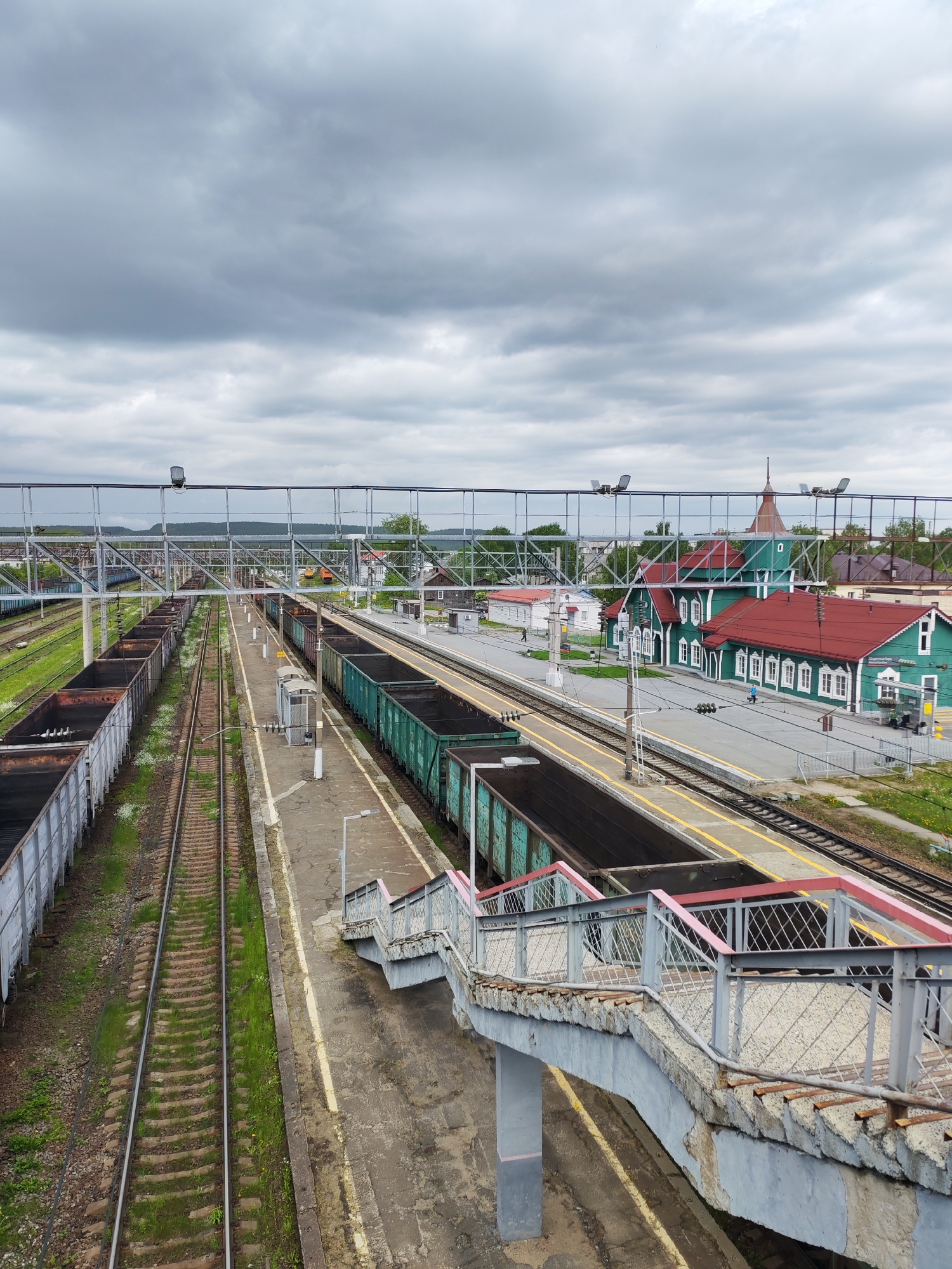 This elevated wide-angle shot captures the railway station in Medvezhyegorsk, Russia, under a heavily overcast sky. The scene is dominated by multiple parallel train tracks, several freight trains, and a distinctive station building.
In the middle and left ground, numerous railway tracks stretch into the distance. A long line of open-top, light-grey freight wagons is parked on the outermost left track. Another set of tracks to the right holds green open-top freight cars, positioned alongside concrete platforms marked with yellow safety lines. Green grass grows between some of the tracks and along their edges.
A robust, grey metal overhead structure, likely supporting railway electrification or signals, spans across the tracks, fitted with various cables and lights.
On the right side of the image, beyond the platforms, stands a prominent two-story station building. It features a bright green facade, multiple red gabled roofs, and white window frames and trim. One section of the roof includes a small, pointed turret-like spire. A sign above the main entrance, though difficult to read clearly, appears to display "МЕДВЕЖЬЕГОРСК," confirming the location. Two small, indistinct human figures can be seen near the station building and on the platform.
In the immediate foreground, a concrete pedestrian overpass descends with a set of stairs. These stairs feature weathered grey metal railings with a reddish-pink top bar, showing signs of age and wear. The concrete structure of the overpass also exhibits some deterioration.
The sky is filled with dense, grey clouds, casting a diffused, subdued light over the entire scene. The overall impression is one of a quiet, functional railway hub on a cloudy day.