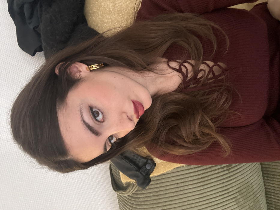 A woman lies sideways on a textured, olive-green surface. She wears a burgundy sweater with criss-cross detailing at the neckline. Her brown hair is styled with loose waves. She has fair skin, defined eyebrows, and dark red lipstick. A gold earring with writing on it is visible in her left ear. Part of a black coat is visible on the left. She looks directly at the camera.