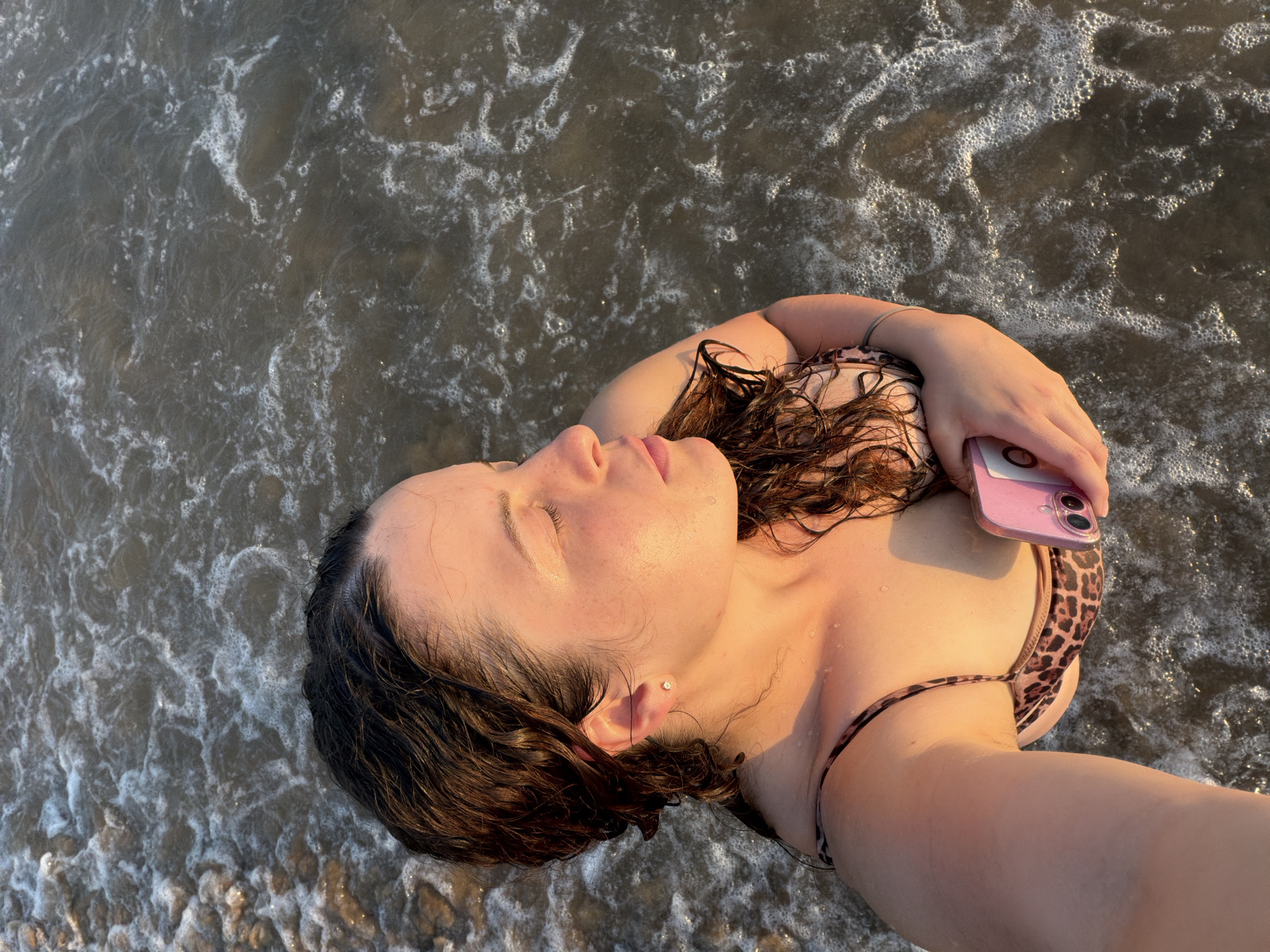 A young woman with wet, dark hair stands in shallow ocean water, taking a selfie with a pink smartphone. She is wearing a leopard-print bikini and a small stud earring. The sun is setting, casting a warm glow on her face and the water. The sea is calm, with small waves lapping at her body. The background consists of the ocean and the shore. The image is taken from a high angle, looking down at the woman. It appears to be taken on a beach in Manavgat, Turkey, given the warm, sunny weather and the presence of the sea.