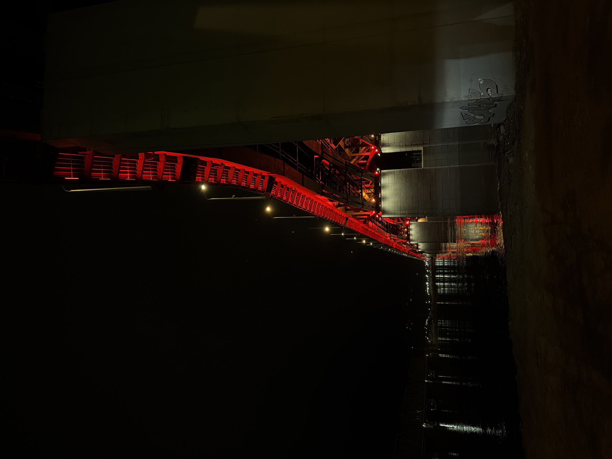 This nighttime image shows the illuminated underside of a bridge in Krasnoyarsk, Russia. The bridge structure is predominantly red with regularly spaced lights along its length. The bridge's reflection is faintly visible in the dark water below. To the right, a concrete support structure and the riverbank are visible, while the left side is mostly black, suggesting deep water. A white graffiti-like drawing can be seen on the concrete under the structure.