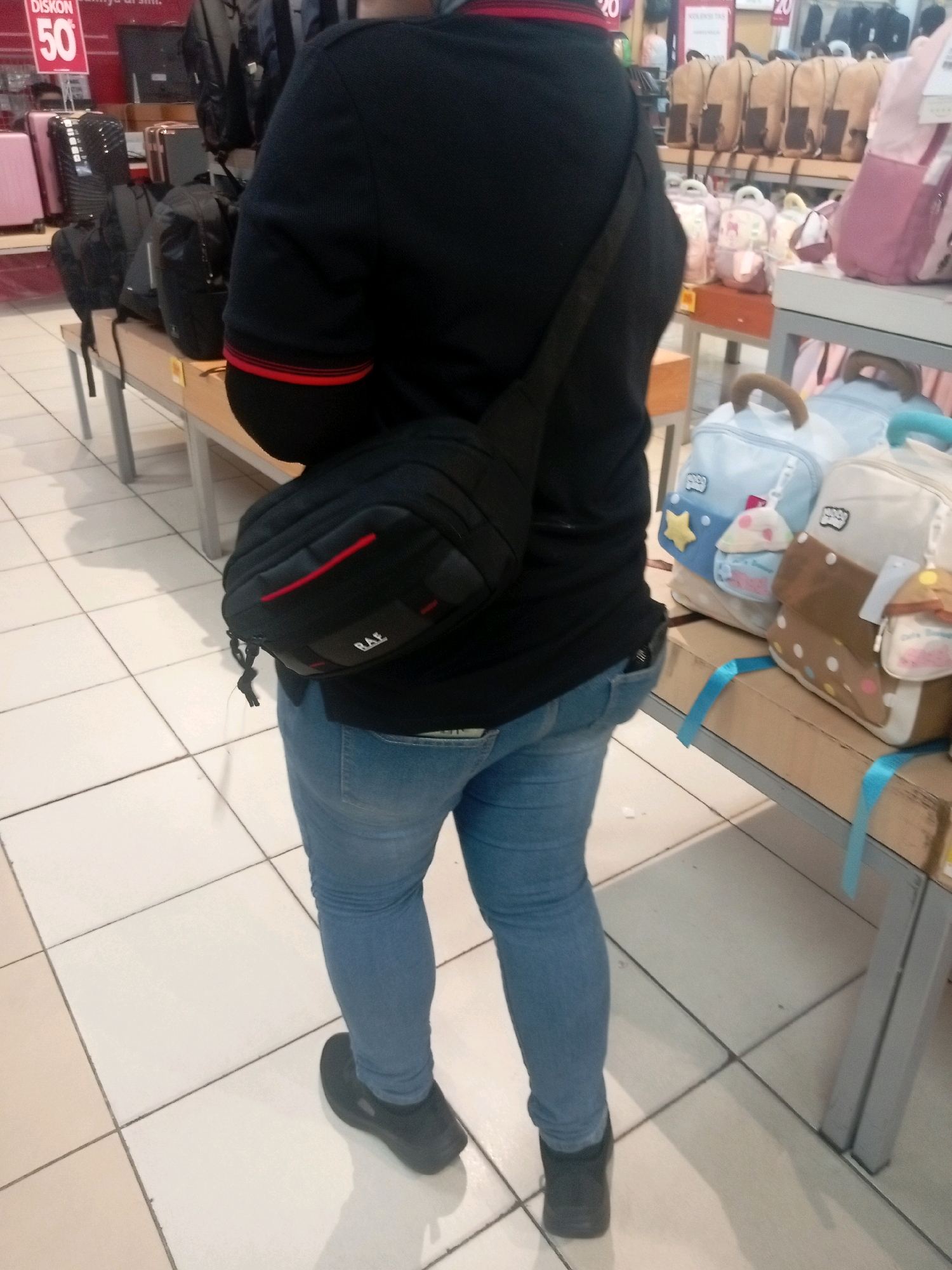 A person is standing on a tiled floor in what appears to be a retail store. The person is wearing a black long-sleeved shirt with red trim on the collar and cuffs, light blue denim jeans, and black slip-on shoes. They are carrying a black cross-body bag with red accents.
In the background, shelves are visible displaying various bags and luggage. To the left, pink suitcases are on display. Further back, a red sign with "DISK. 50" is prominent.
The environment suggests a shopping area, likely a department store or a specialty bag shop. Based on visual cues of the products and general store aesthetic, this scene is consistent with a retail location in Batam, Indonesia.