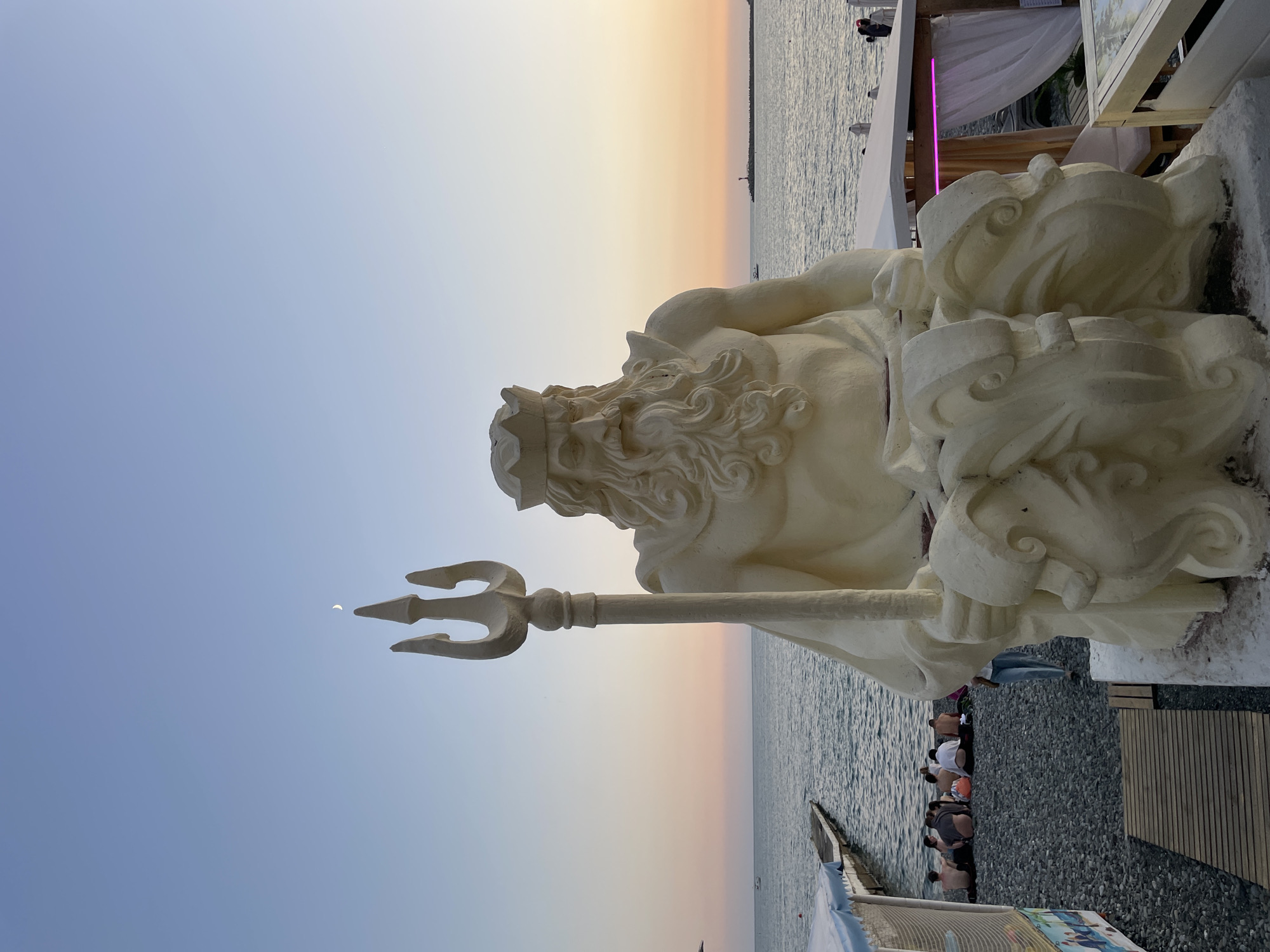 An imposing statue of Neptune, the Roman god of the sea, dominates the foreground of the image. He is depicted as a muscular figure with a long beard and flowing hair, wearing a crown and holding a trident. The statue is a light, creamy color and appears to be made of concrete or a similar material. It is seated on a decorative base.
The statue is positioned on a pebble beach, with the sea stretching out behind it. The water is calm, reflecting the soft colors of the sunset. A wooden pier extends into the sea on the left side of the image, and several people are seen sitting on the beach near it, facing the water.
In the background, a lighthouse stands on the horizon, and a small boat is visible on the sea. The sky is a gradient of soft blues and pinks, indicative of twilight. To the right of the statue, there are white cabanas with pink lighting, suggesting a beachside recreational area.
This scene is located on the Black Sea coast in Sochi, Russia, likely in the Adler district, a popular resort area known for its beaches and statues. The overall atmosphere is serene and picturesque, capturing a moment of tranquility at the end of the day.