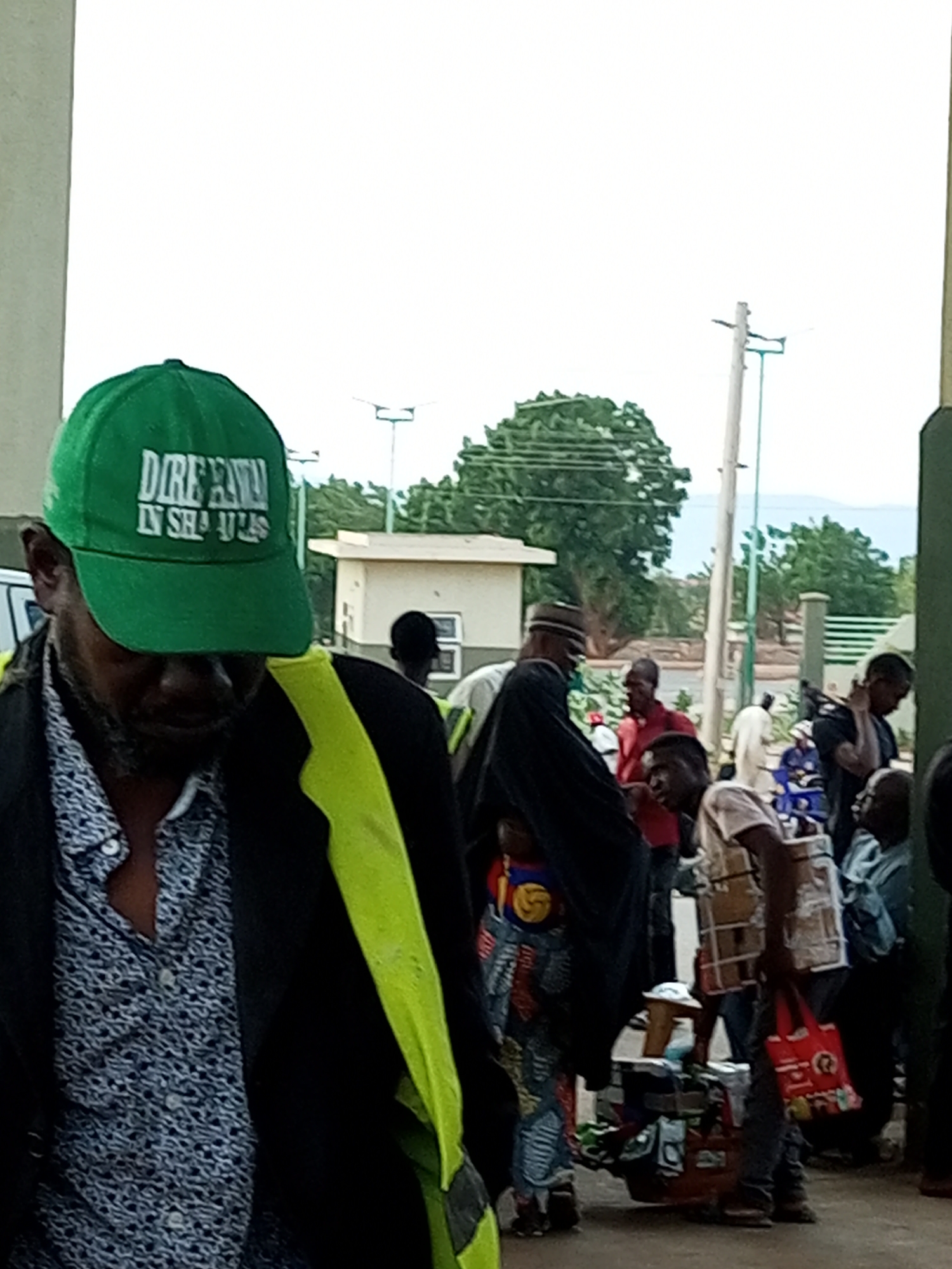 The image depicts a street scene in Gombe, Nigeria, likely near a market or transport hub. In the foreground, a man wearing a green baseball cap with white lettering and a patterned shirt under a black jacket with a neon yellow accent is partially visible. The lettering on his cap appears to read "DIREKA" and "IN SHA ULAS," though the latter is partially obscured. He is looking down, and his beard is visible.
Behind him, several other individuals are present. To the left, a small section of a white vehicle is visible. Further back, there are buildings, including a low-rise structure with windows and a tiled roof. Trees and utility poles are scattered throughout the background, suggesting an outdoor setting. The overall atmosphere is that of a bustling public space with people engaged in various activities. Some individuals are carrying bags or merchandise, hinting at commerce or travel. The lighting suggests it is daytime.