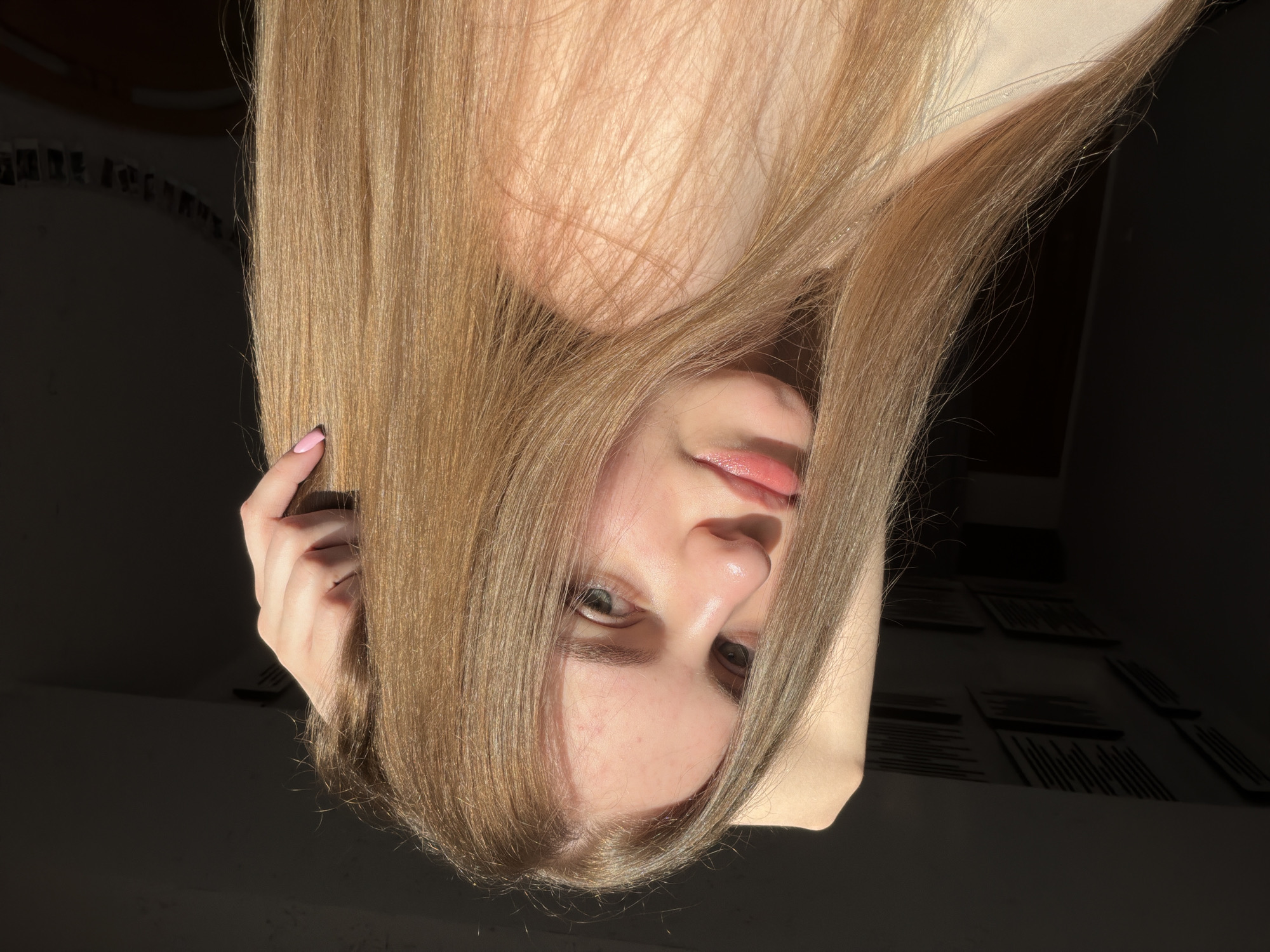 A young female with long, light brown hair is captured in a close-up portrait. She is holding her right arm behind her head, with her hand touching her hair. Sunlight illuminates her face, casting a shadow on the right side of the frame. She is wearing a light-colored top. The background is dark and appears to be an indoor setting, possibly a room with some textured patterns on the ceiling. There are no visible clues to suggest the location is the city of Dnipro, Ukraine.