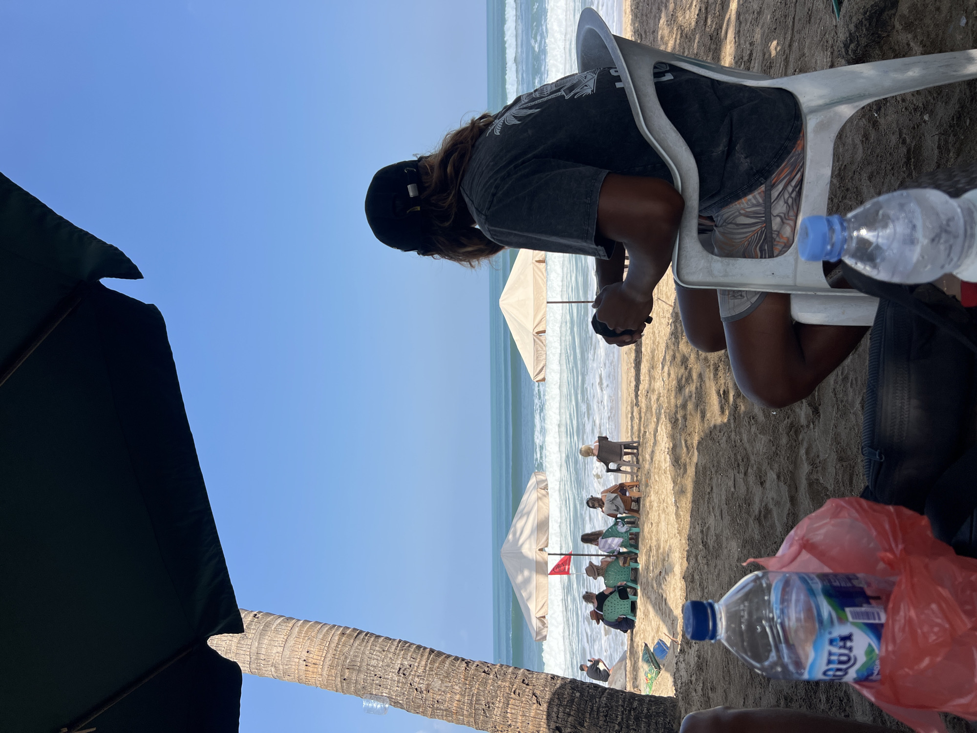 In Kuta, Indonesia, a man wearing a black baseball cap and a dark t-shirt with a graphic print sits facing the ocean in a white plastic chair on a sandy beach. He has long, dark, wavy hair and is wearing patterned shorts. His hands are resting in his lap, and he appears to be holding a black object, possibly a camera or phone. In the foreground, a bottle of Aqua brand water is placed inside a red plastic bag. Another partially visible water bottle with a blue cap is on the sand in front of the man.
In the middle ground, several people are seated on chairs under a large, light-colored umbrella on the beach, facing the ocean. Another umbrella is visible further down the beach. The ocean has moderate waves, and the sky is clear and blue. A palm tree trunk is visible on the left side of the frame, partially obscured by a large dark umbrella overhead. The overall scene depicts a relaxed atmosphere on a sunny day at the beach.