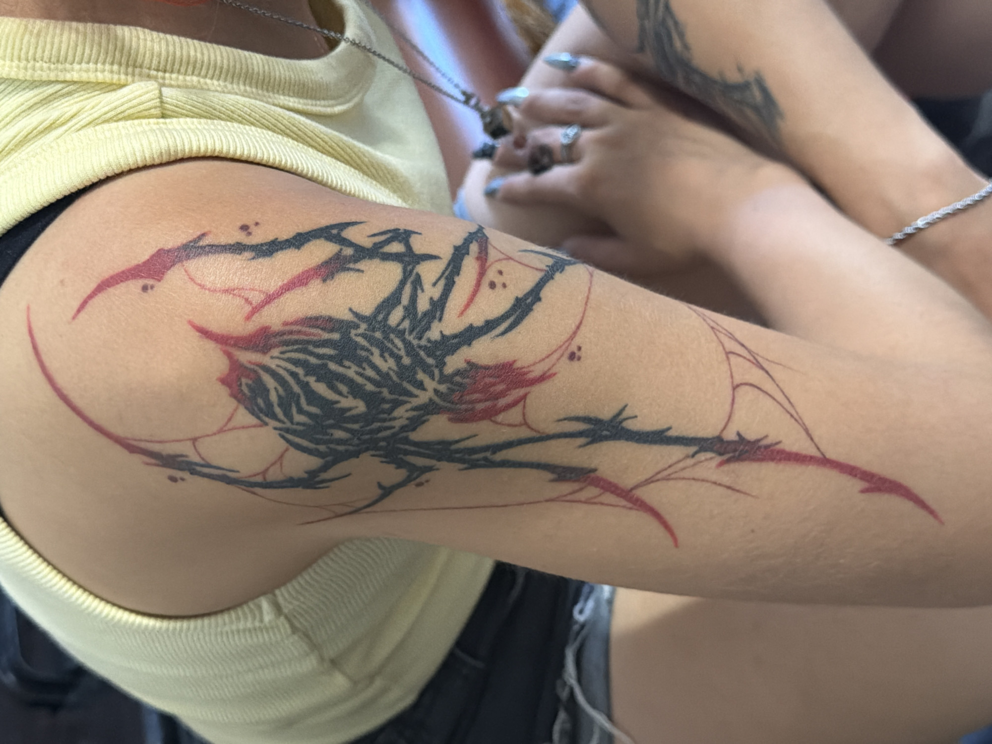 The image shows a close-up of a person's arm with a black and red spider tattoo. The tattoo is abstract and stylized, with sharp lines and a tribal aesthetic. The person is wearing a yellow ribbed tank top and denim shorts. Their hands are visible in the foreground, with manicured nails and rings on their fingers. The background is blurred, but suggests an indoor setting. There are no discernible landmarks or features that would indicate the city of Hostalric, Spain.