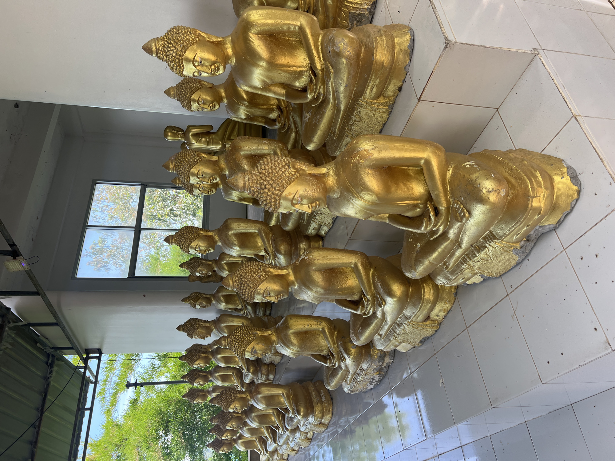 A row of gilded Buddha statues sits on tiled platforms under a covered walkway. The statues are in a seated meditation pose, with their hands resting in their laps. In the background, a window looks out onto lush green trees and a bright blue sky. The scene is peaceful and serene, suggesting a place of worship or contemplation. The image appears to be taken in Baan Thong Krut, Thailand, a location known for its tranquil atmosphere and Buddhist temples.
