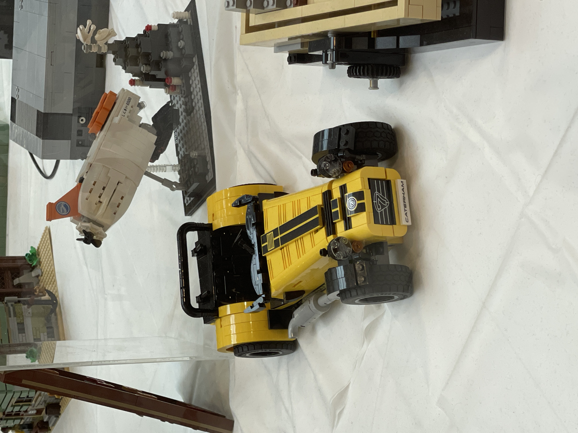 The image displays a collection of LEGO models, including a yellow Caterham 7 car prominently in the foreground. The car is detailed with black racing stripes on the hood, a roll bar, and a black interior. To the left of the car is a clear acrylic display case containing what appears to be a LEGO building with a brown facade. Behind the car, several other LEGO creations are visible, including a beige and orange spacecraft on a stand, a dark grey LEGO structure resembling a console, and a jagged black rock formation with red accents. To the right of the Caterham car, a portion of another LEGO structure, primarily tan in color, is visible. The models are arranged on a white surface, likely a table or display cloth, suggesting an indoor exhibition or event. The environment is well-lit, and the focus is on the LEGO models, with the background slightly blurred. There are no people visible in the image. The image was taken in Sofia, Bulgaria.