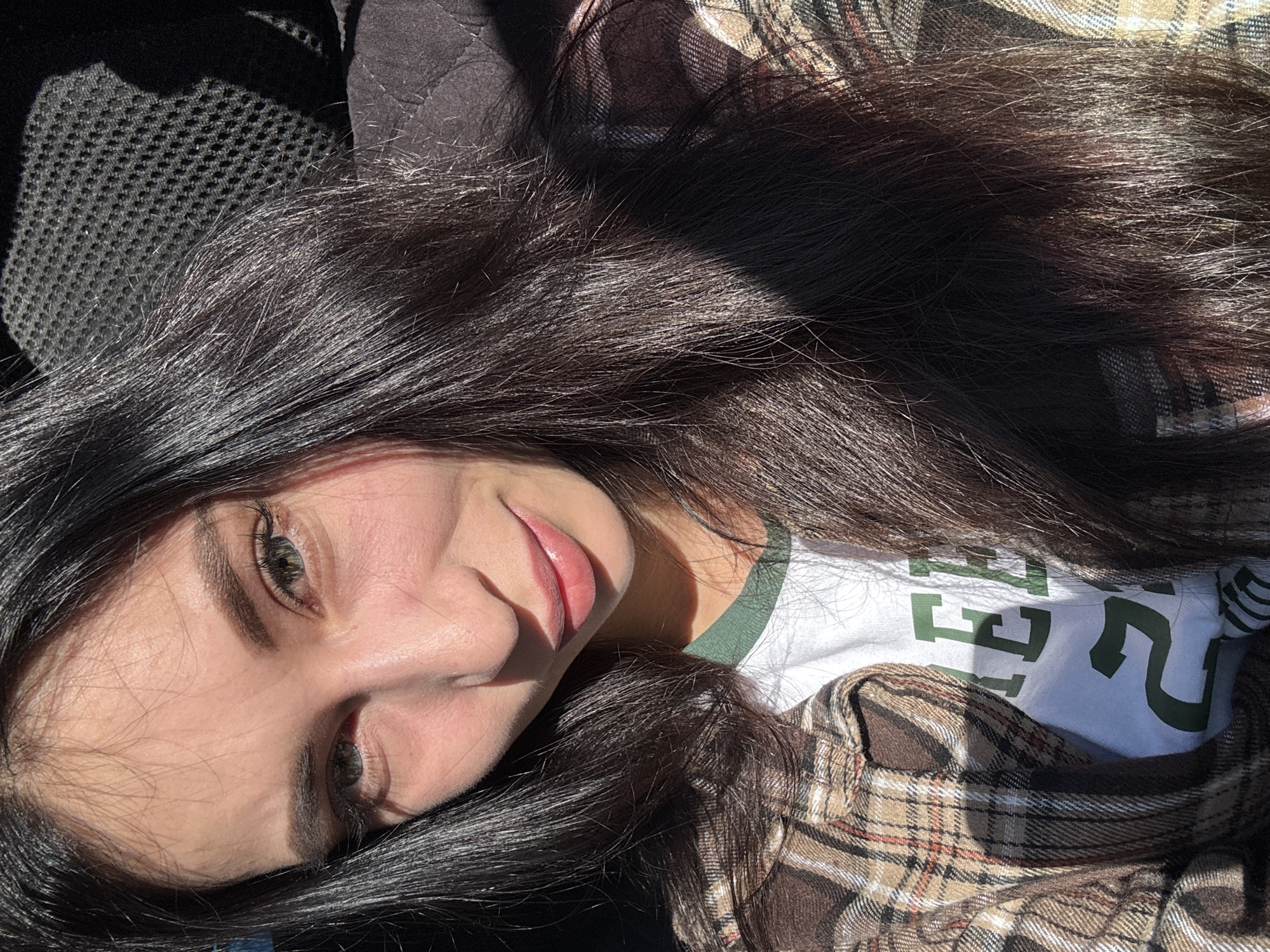 A young woman with long, dark hair and light eyes is lying down, looking at the camera. She is wearing a white T-shirt with green lettering and a brown and beige plaid flannel shirt. Her face is partially in shadow. The background appears to be a car interior with a black seat. The location in the city of Chaikovsky, Russia, cannot be determined from the image.