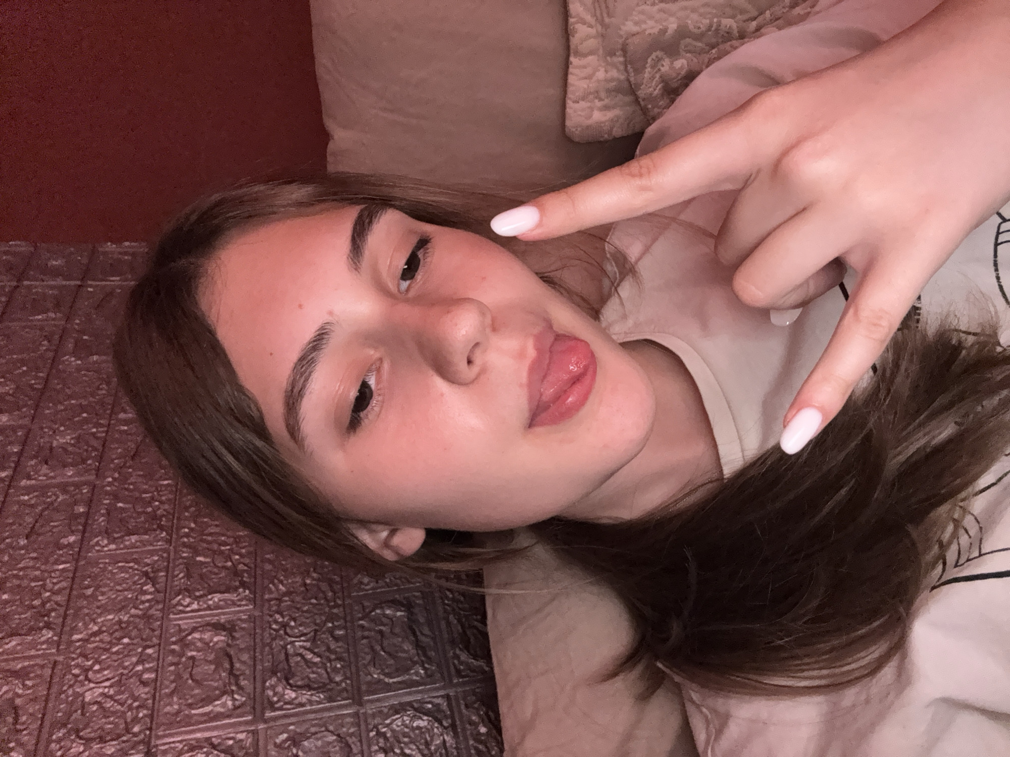 The image features a young woman lying down, captured in a close-up, angled shot. She looks directly at the camera with a playful expression, her tongue slightly extended. Her left hand is raised, making a "rock on" gesture with her index and pinky fingers extended, while her other fingers are folded. Her fingernails are painted a glossy white. She has dark brown hair that cascades around her face and over her shoulders.
The setting appears to be indoors, with a textured, dark reddish-brown surface visible on the left side of the frame, possibly a wall or headboard. On the right and beneath her, there's a light beige or off-white pillow or cushion and a soft, light-colored fabric, possibly part of a bed or sofa, with a subtle pattern. The lighting is warm and soft, suggesting an indoor environment, potentially in the evening. The overall mood is casual and lighthearted. No text is visible in the image. The location is Panagyurishte, Bulgaria.