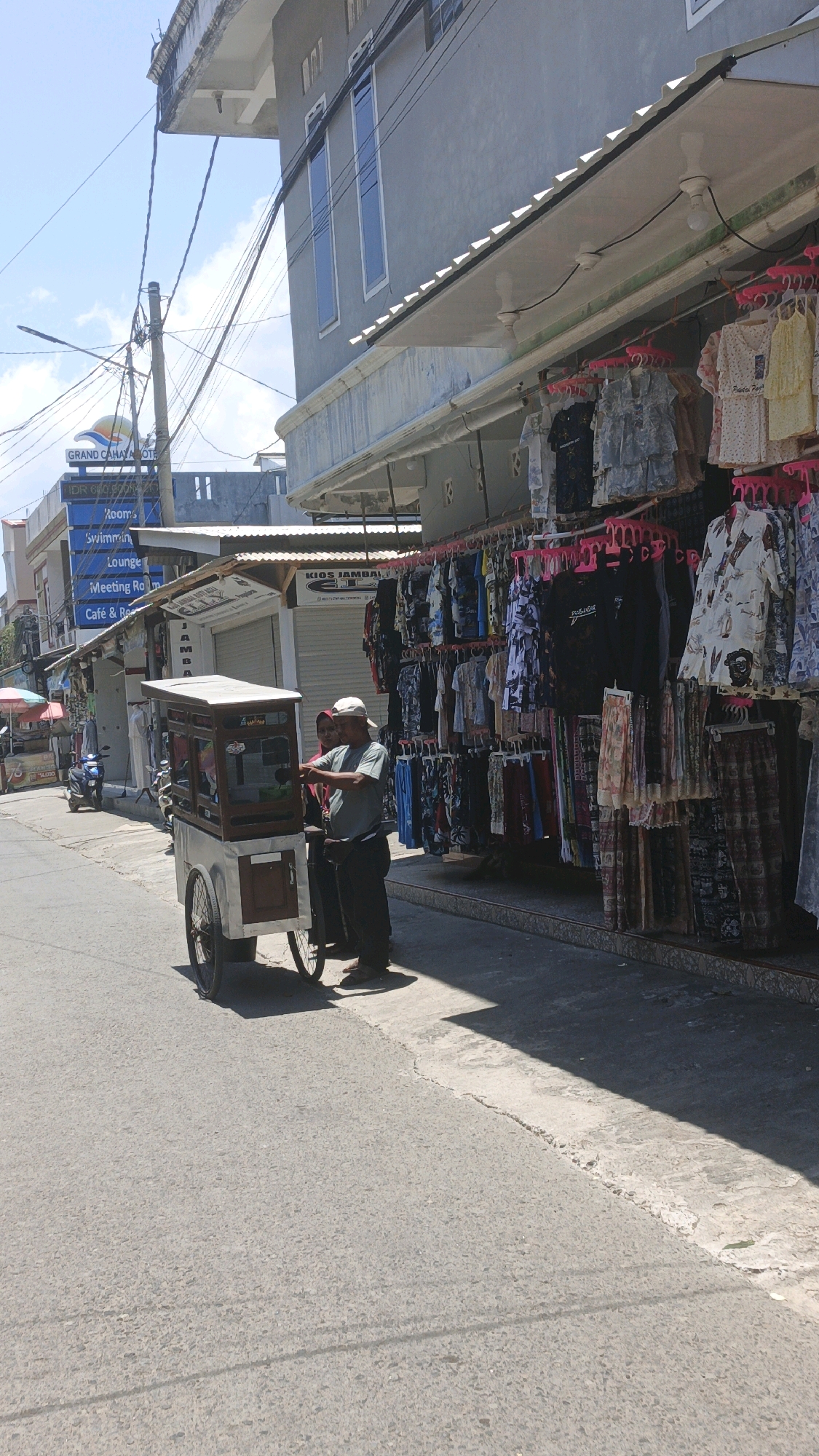 In Desa Pagandaran, Indonesia, a man in a white cap and grey shirt operates a mobile food cart with a glass display on a paved road. The cart has a wooden top and a silver and white metal base with two spoked wheels. Beside the cart, a woman in a red headscarf and dark clothing is visible. The street is lined with shops displaying clothing on racks under awnings. The shops have signage, with one appearing to be "KIOS JAMBALY". The buildings are two to three stories tall with a gray facade and windows. Overhead, electrical wires crisscross against a clear blue sky. The scene is illuminated by bright sunlight, casting shadows on the road. In the background, a blue signpost indicates "GRAND CAHAYA HOTEL", "Rooms", "Swimming", "Lounge", "Meeting Room", and "Cafe & Restaurant". A motorcycle is parked on the left side of the street.