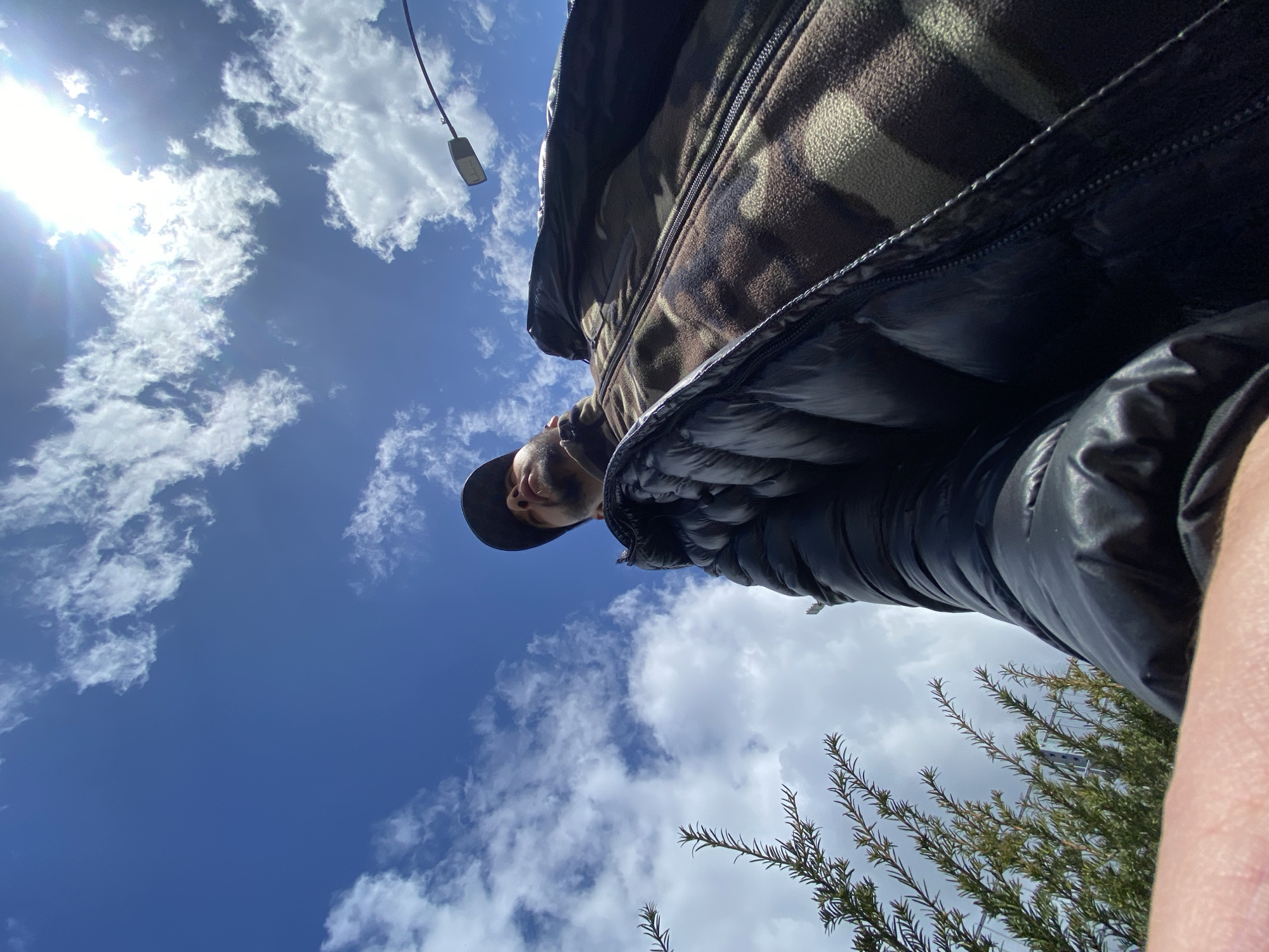 A low-angle shot reveals a person wearing a camouflage fleece and a black jacket. The person also appears to be wearing a hat. Above the person, the sky is blue with scattered white clouds, and the sun is visible, appearing very bright. A streetlight is visible in the upper left corner of the image. Some green foliage is visible in the lower right corner. This image was taken in Bischheim, France.