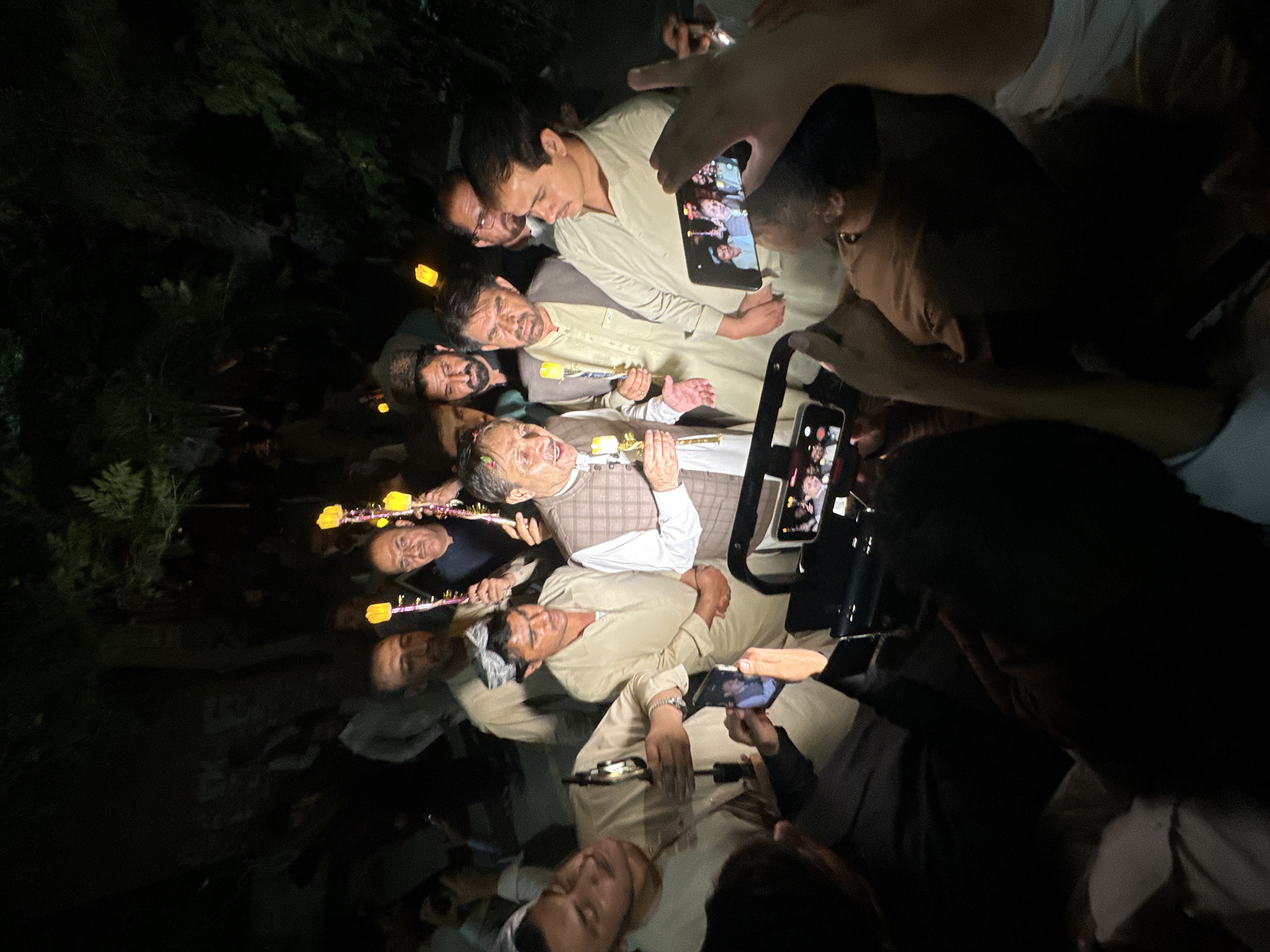 In Quetta City Tehsil, Pakistan, an outdoor gathering takes place at night, illuminated by the glow of several lit candles held by individuals. A man, distinguished by a plaid vest over a white shirt and a traditional cap, stands at the center, holding a candle and appearing to speak or sing. He is surrounded by a crowd of men, many of whom are dressed in traditional Pakistani attire. Several people are using their mobile phones to capture photos or videos of the event. The background consists of dark foliage, suggesting the presence of trees or bushes. The overall scene depicts a celebratory or commemorative event.