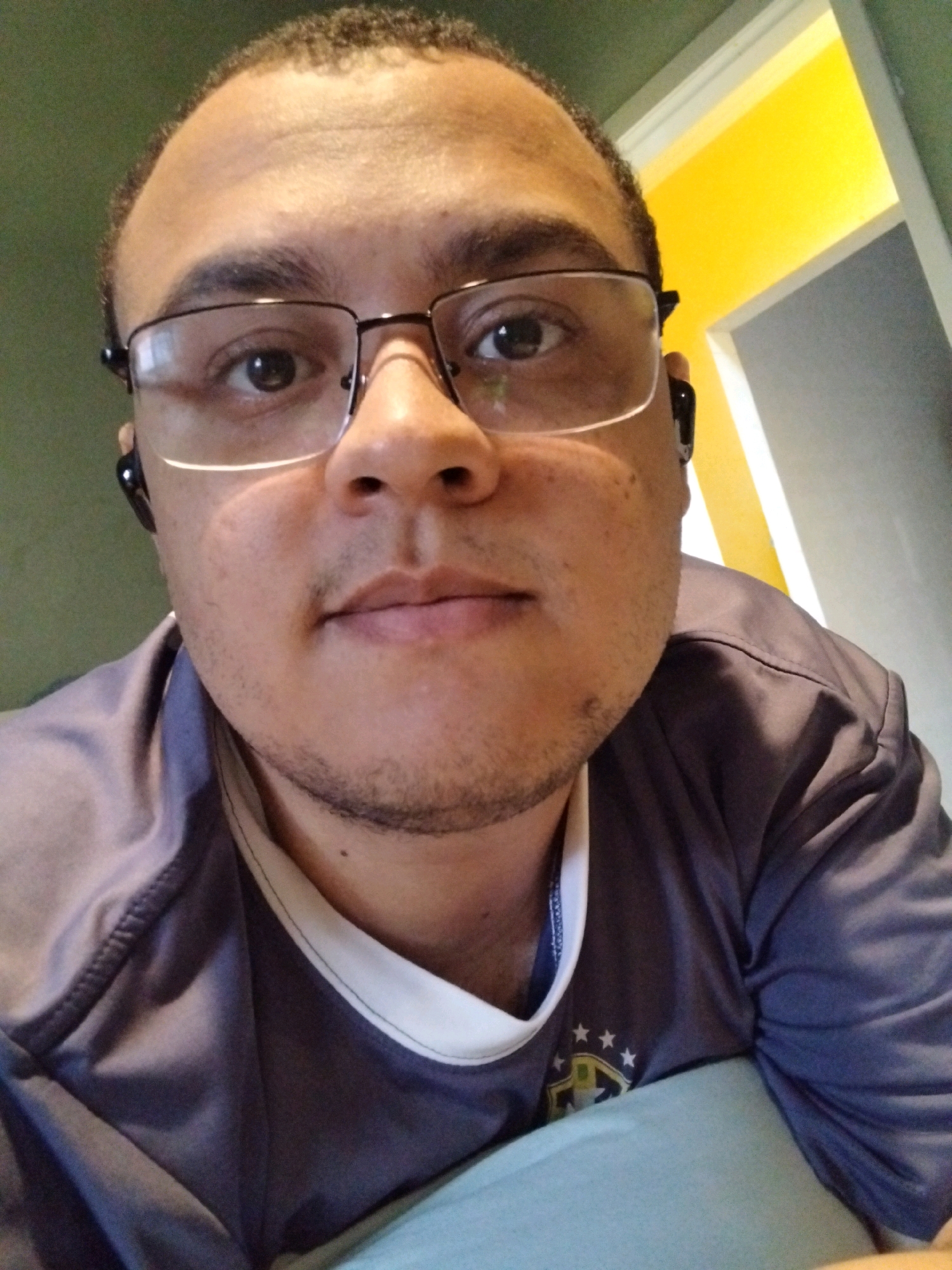 A man with dark skin, wearing glasses and black earbuds, is looking at the camera. He has short, curly dark hair and light stubble on his chin. He is wearing a dark purple shirt with a white collar and a crest with stars on it. The background is blurry, with a green wall on the left and a yellow and white wall on the right, possibly indicating an indoor setting. The image appears to be a selfie. The scene is likely in the city of São João De Meriti, Brazil, as per the prompt.