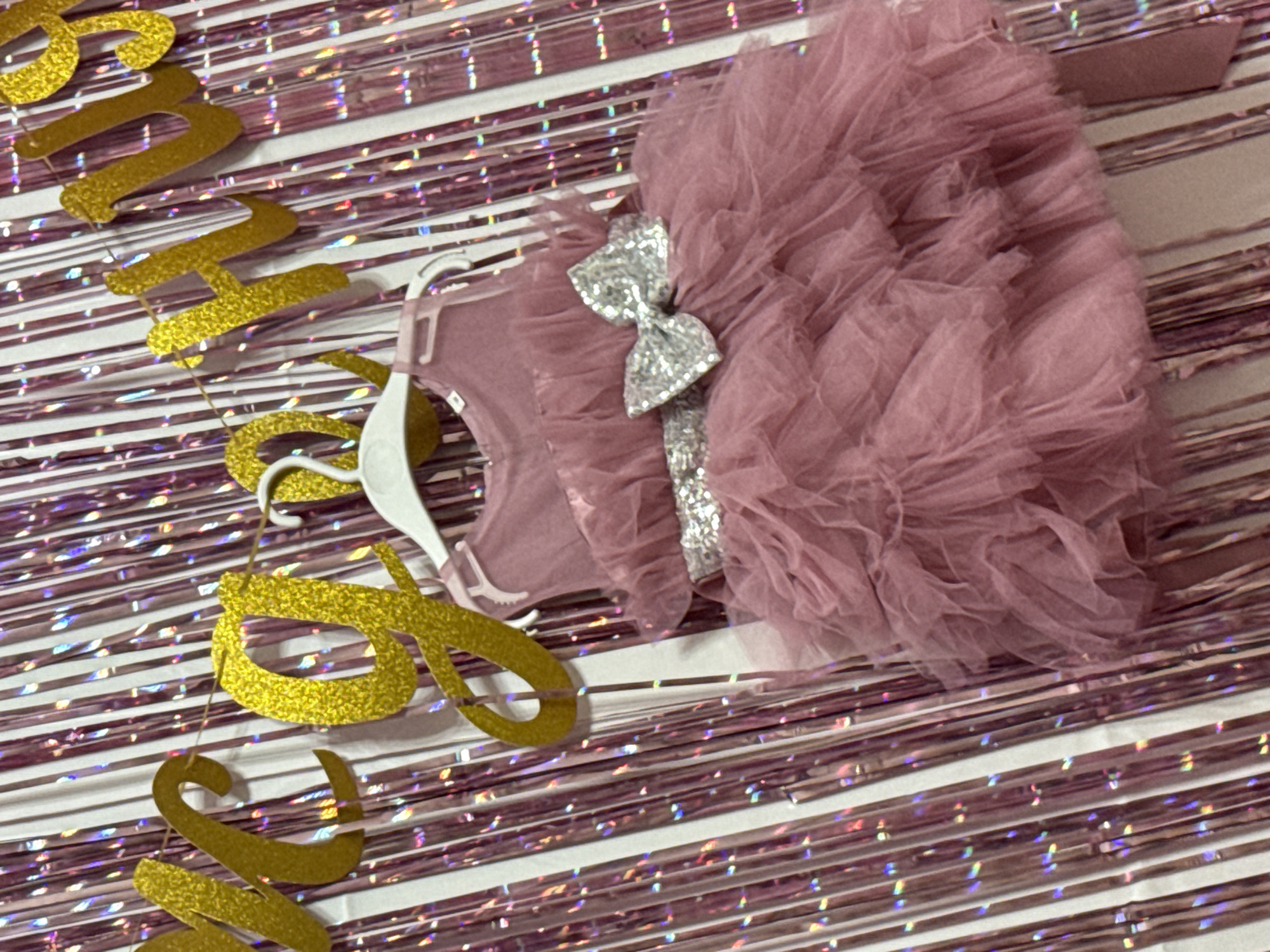 A dusty rose, ruffled tulle dress with a silver sequined bow and belt hangs from a white hanger. The dress is displayed in front of a backdrop of pink, holographic tinsel fringe and a gold glitter banner that reads "It's a Girl". The scene appears to be set up for a baby shower or a celebratory event. The image is taken indoors.
