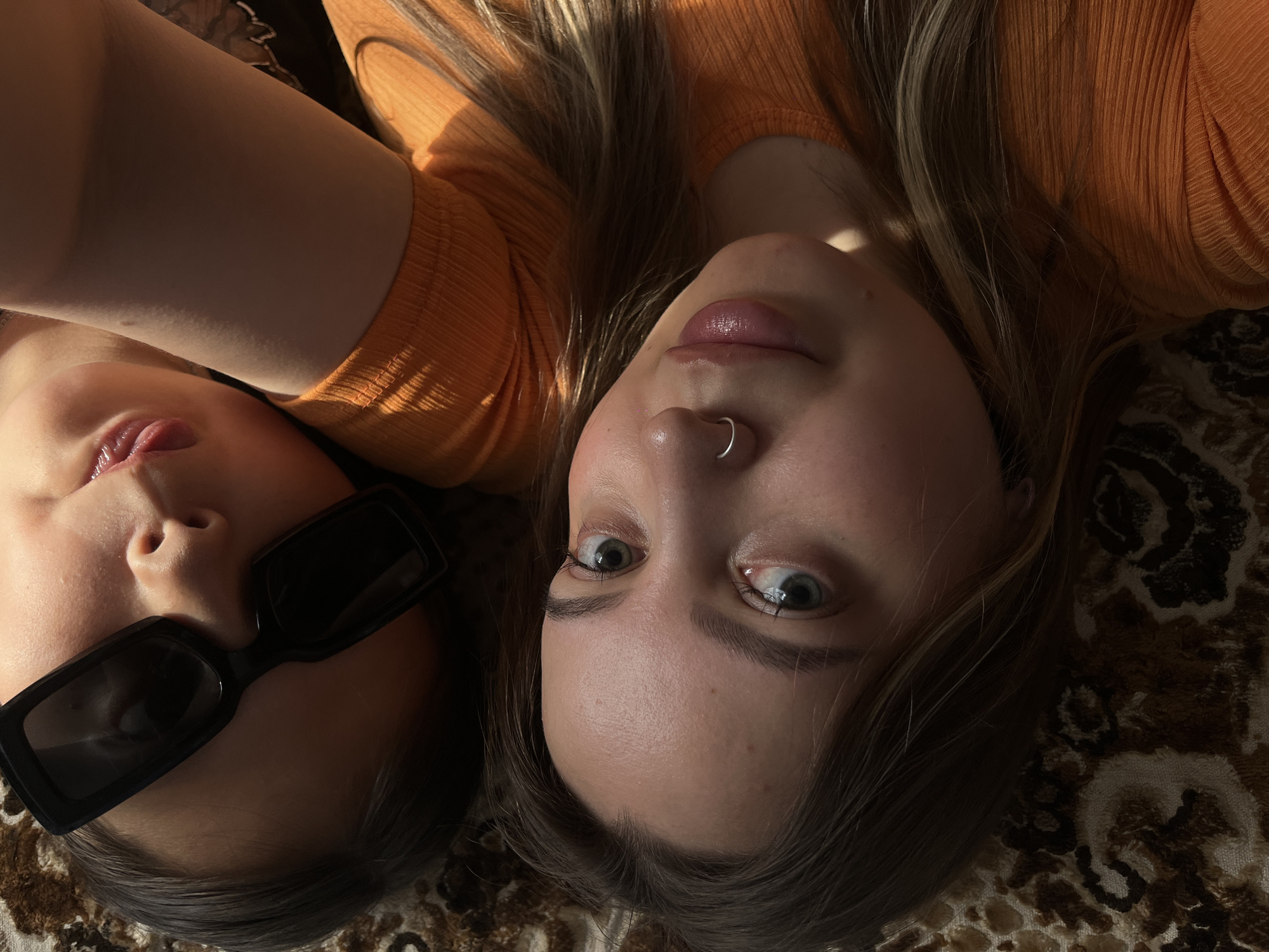 Two young women are lying down, viewed from above, with their heads close together. The woman on the right has light brown hair, fair skin, and green eyes. She is wearing an orange, ribbed top. She has a small silver hoop nose ring and is looking directly at the camera with wide eyes, her mouth slightly open. The woman on the left has darker hair and is wearing black sunglasses. She has fair skin and her tongue is slightly sticking out. She is also wearing an orange top. They are lying on a patterned rug. The lighting suggests it is daytime, with sunlight casting shadows and highlights on their faces and hair. The scene appears to be indoors.