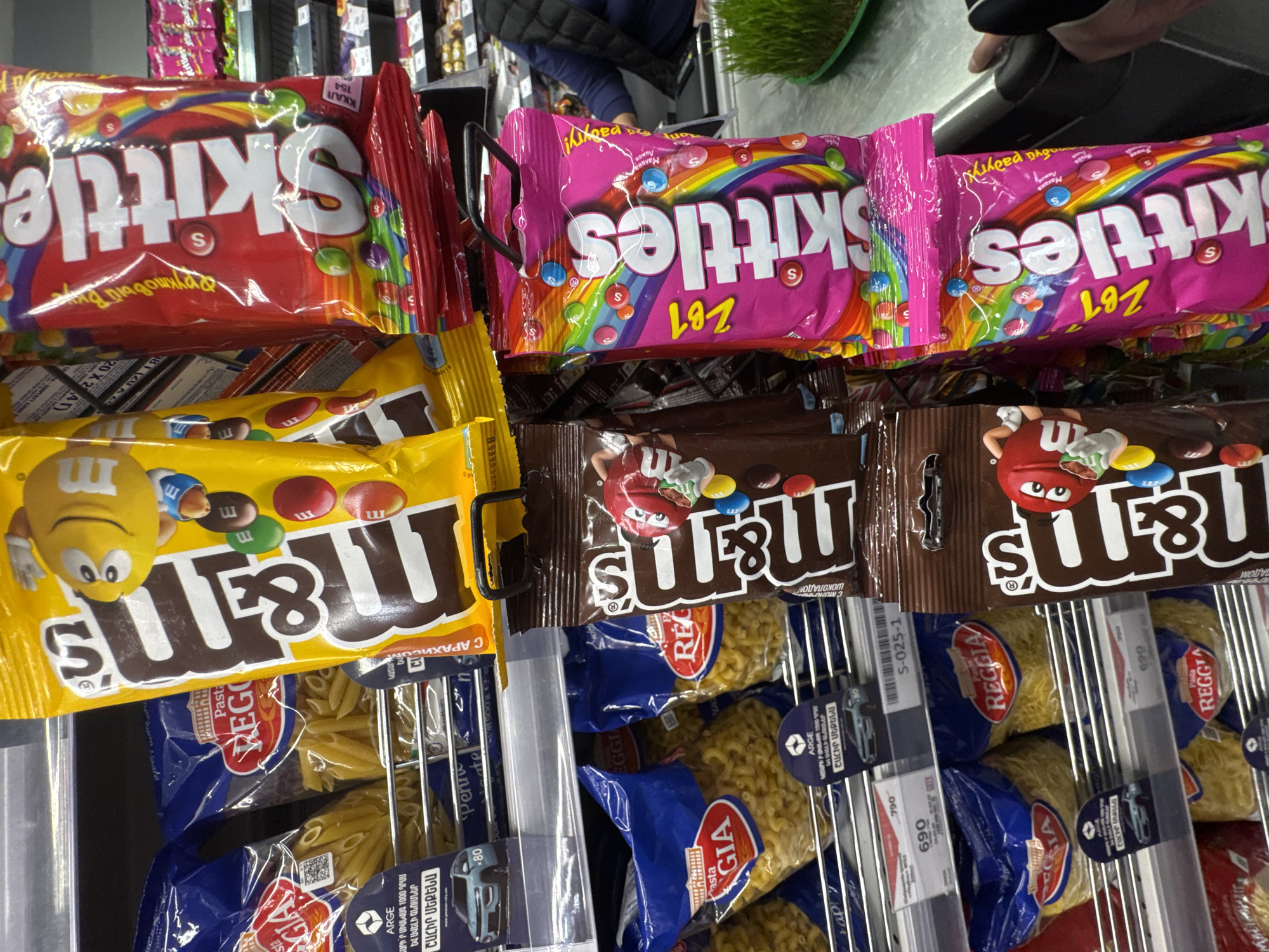 This image appears to be taken in a supermarket, most likely in the candy and pasta aisle. The main focus is on a variety of candy, including Skittles and M&M's, positioned above packages of pasta. Brands of pasta include "Reggia" and "Costa". There are price tags visible for the pasta. The location appears to be in Unknown, Unknown, as indicated by the text "ARGE" and Armenian words on the price tags.
