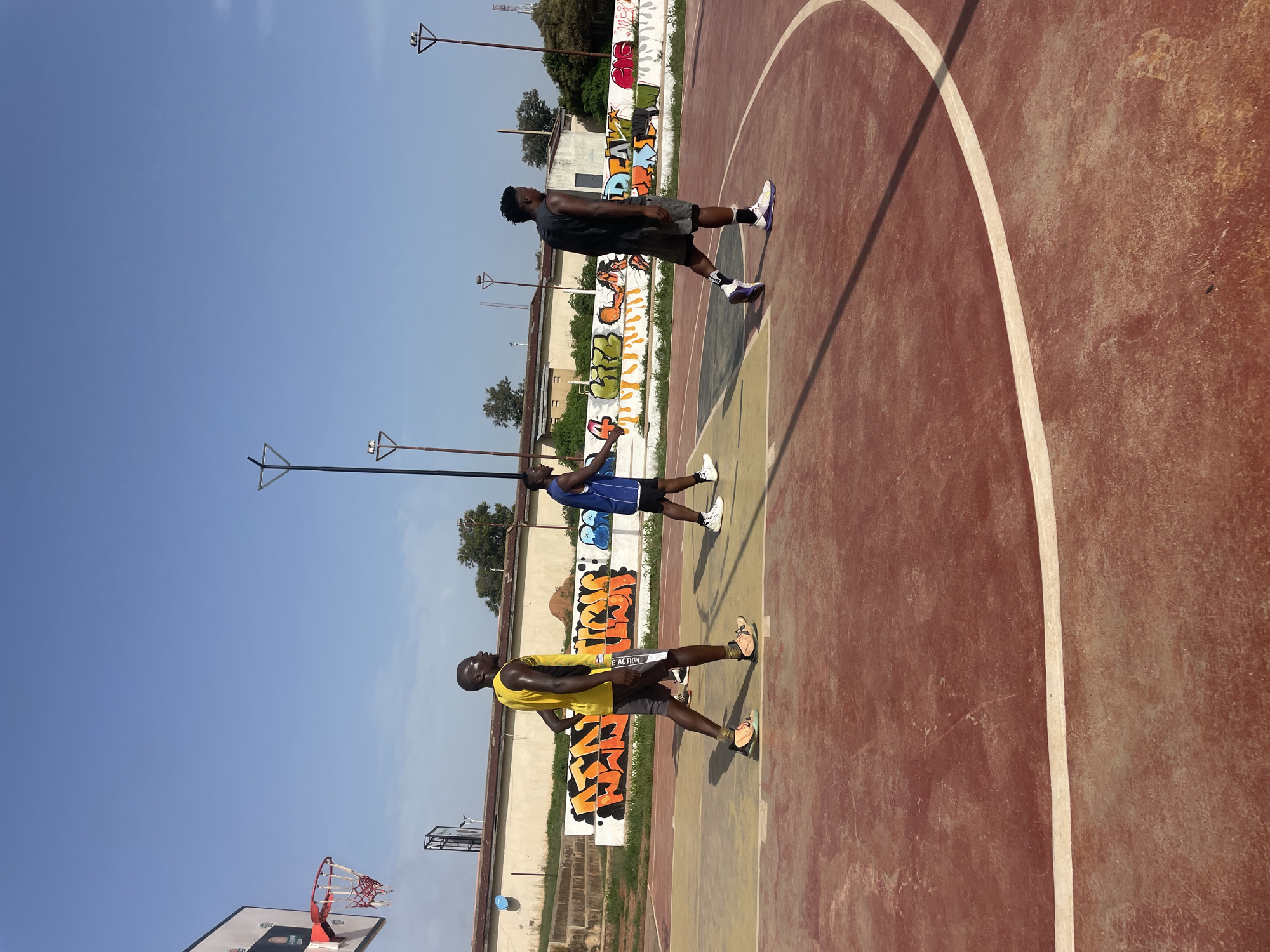 Three men in athletic wear stand on a cracked, outdoor basketball court in Jos, Nigeria. Graffiti adorns a low wall adjacent to the court. Light poles stand around the perimeter. A basketball hoop is visible. The sky is blue and clear.