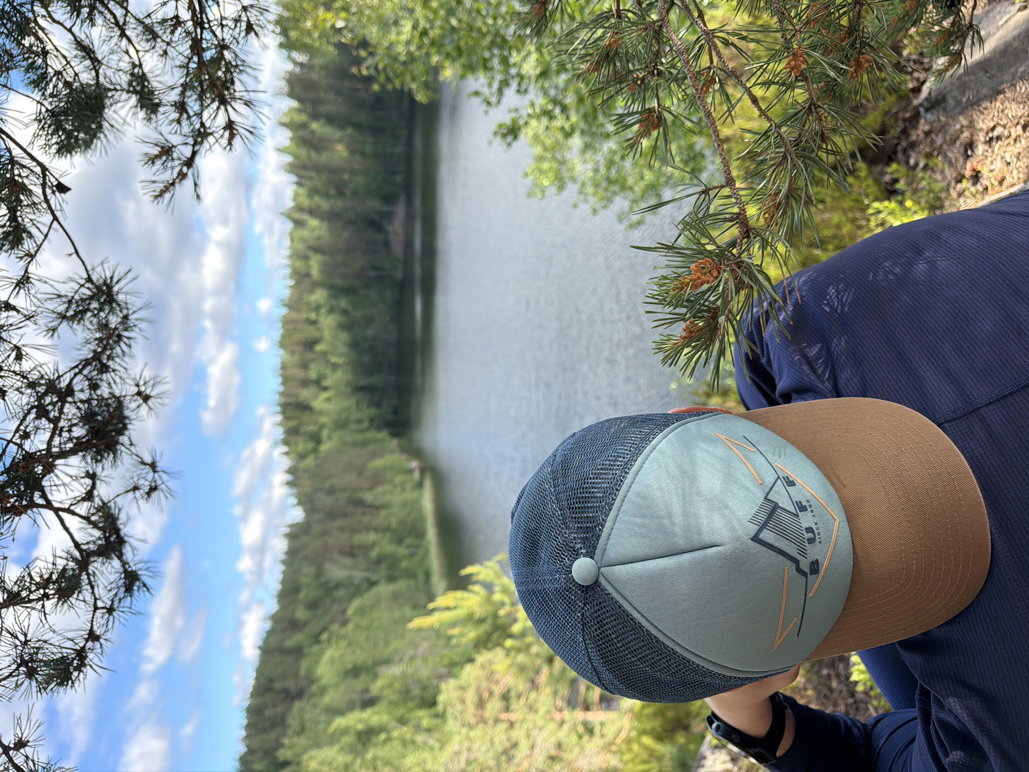 The image shows a person wearing a dark blue shirt and a light blue and brown cap, looking out at a body of water. The person is positioned in the foreground, with their back to the camera, leaning forward slightly. The cap has a mesh back and a light blue front panel with a logo that appears to be "BUFF SINCE 1992" along with a mountain graphic. The background features a serene lake surrounded by a dense forest of green trees. The sky above is blue with scattered white clouds. Pine branches with small, developing cones are in the foreground, framing the view of the lake and forest. The lighting suggests it is daytime, with sunlight filtering through the trees. The overall scene depicts a moment of quiet contemplation in a natural setting. The location is identified as Ruch'i, Russia.