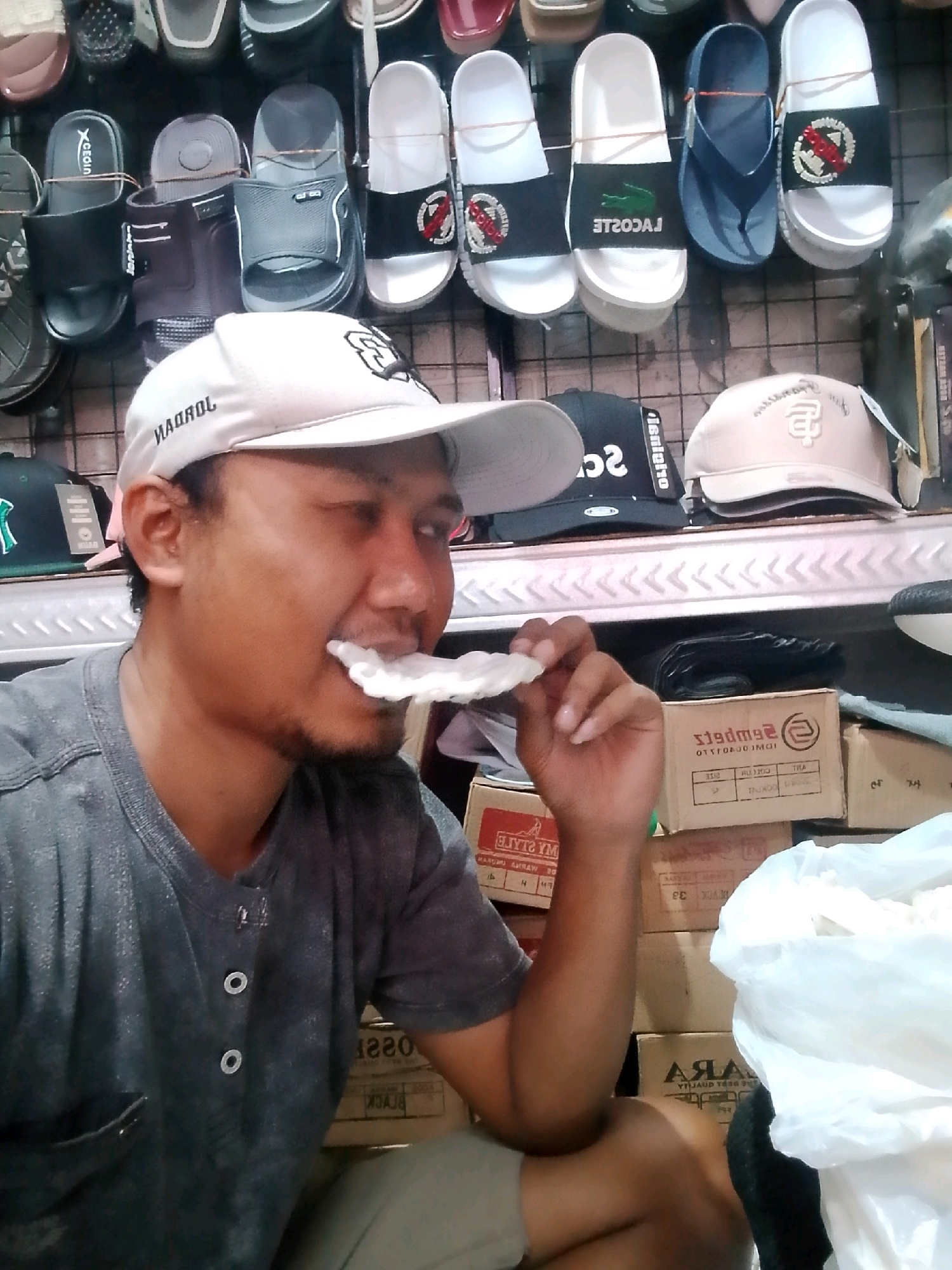 A man wearing a grey t-shirt and a white baseball cap is in the foreground, with his mouth open and a white object, possibly food, held between his teeth. Behind him, shelves are stocked with various footwear, including sandals and flip-flops. Several cardboard boxes are stacked on the floor in the background. The overall setting appears to be a retail establishment, possibly a shoe store, in the city of Sumberjati, Indonesia. The lighting is somewhat dim, creating shadows and highlighting the details of the man and the merchandise.