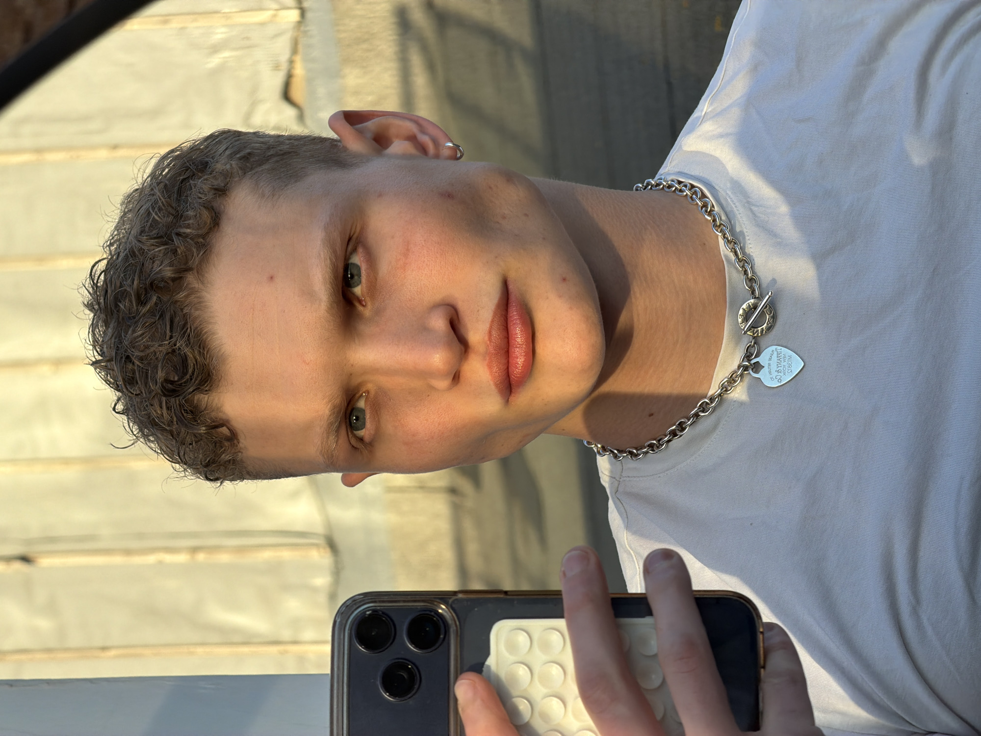 A fair-skinned person is taking a selfie with a dark-colored smartphone. They have short, curly hair, and their eyes are visible. They are wearing a white t-shirt and a silver chain necklace with a heart-shaped pendant. An earring is visible in their left ear. The background appears to be an exterior wall with some horizontal structures, possibly steps. The light is bright and sunny, casting shadows. This information is not sufficient to determine the location in London.
