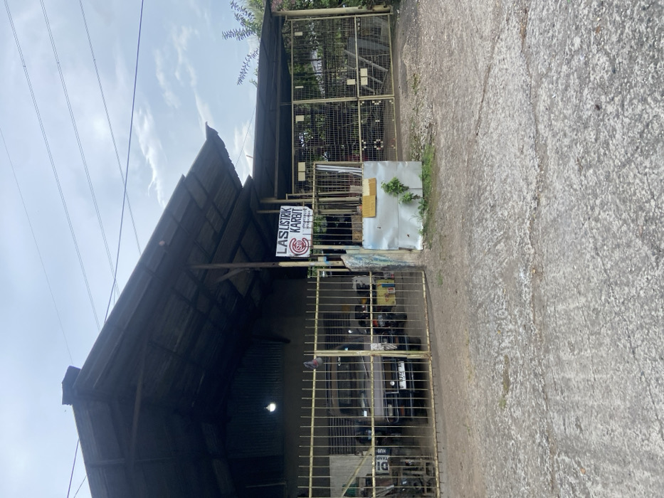 The image shows the exterior of a small, enclosed workshop or garage in Cipatat, Indonesia, located at coordinates -6.788, 107.434. The building is constructed with a corrugated metal roof that extends to the edge of the property and metal bars on its exterior walls. A small, gray vehicle is parked inside the workshop. A sign on the exterior wall reads "LAS LISTRIK KARBIT". There are no people or animals visible in the image. The image appears to have been taken in the early afternoon, on a cloudy day. The ground is covered in loose gravel and dirt, and there is a small amount of vegetation in front of the building. There are power lines running across the sky. The image is slightly out of focus.