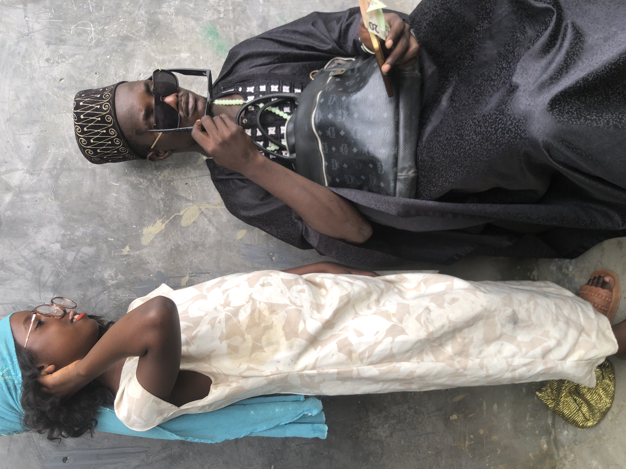 In the city of Ado, Nigeria, a young girl and an adult man are positioned against a textured, gray concrete wall. The girl, on the left, wears a light-colored dress with a subtle floral pattern, a bright blue headscarf, and round, light-rimmed glasses. Her right hand is raised to her ear, and her lips are painted red. Beside her, a man is seated, dressed in a dark, patterned robe and a patterned cap. He holds large, dark sunglasses in his right hand, and wears a green beaded necklace. In his left hand, he holds a wallet with several banknotes visible. A black handbag with a distinctive monogram pattern rests on his lap. The ground beneath them is concrete, with a golden, textured item visible near the girl's feet.