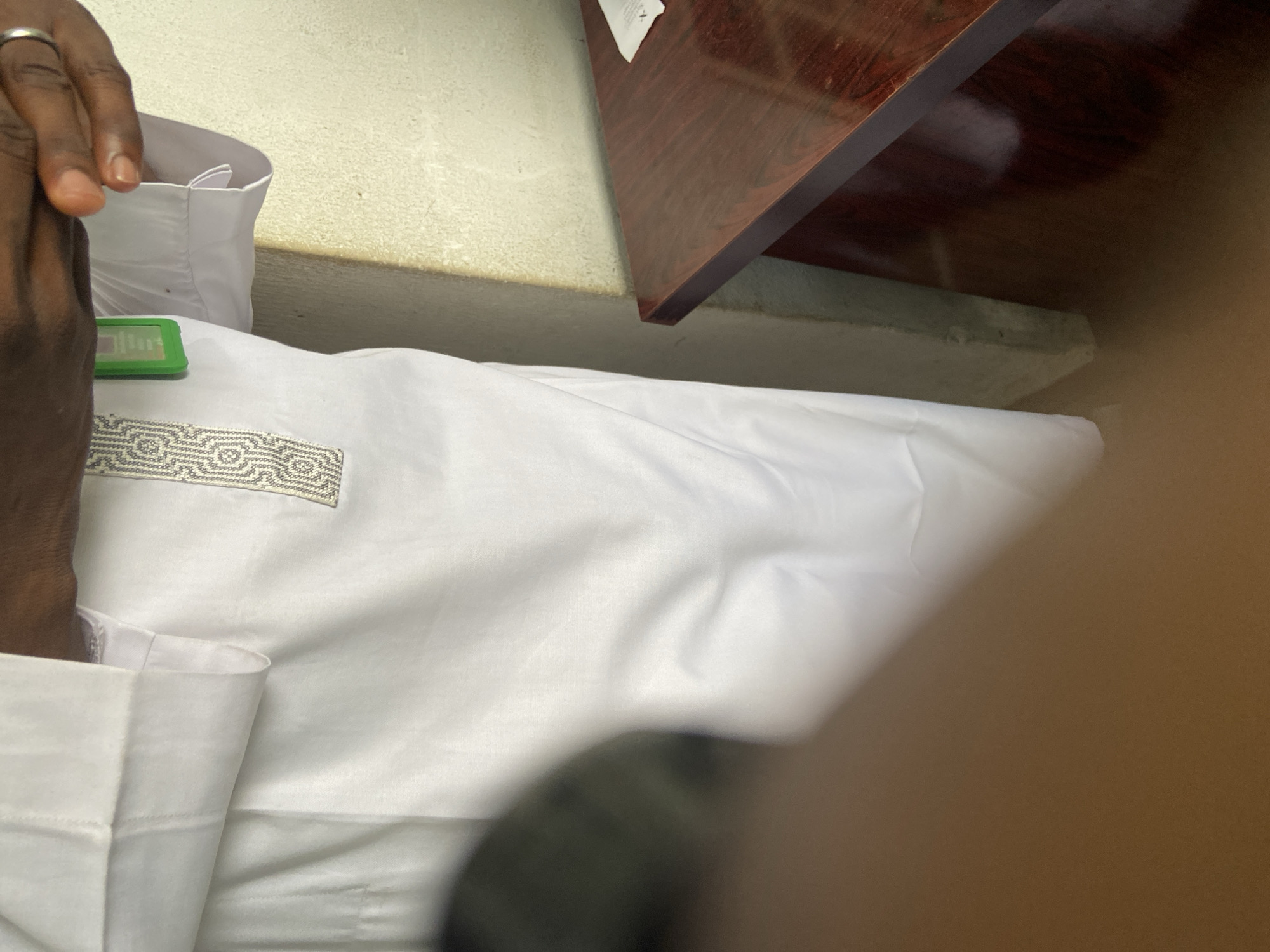 In Kuson, Nigeria, an individual wearing a white thobe with intricate silver embroidery on the chest is standing. Their hands are visible, one of which is adorned with a ring. The thobe has a neatly folded collar. The background shows a light-colored wall and a section of a dark wood desk. A green object, possibly an identification badge, is clipped to the thobe. The lighting suggests an indoor setting.