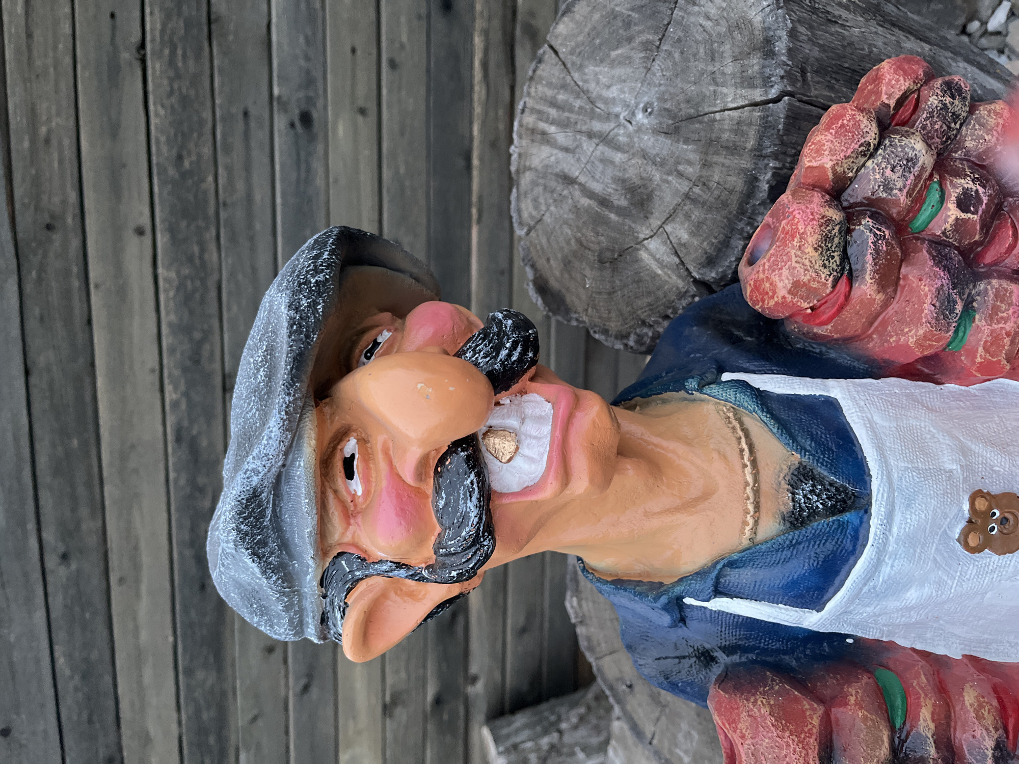 This is a medium shot, eye-level, outdoor photograph of a resin garden statue of a caricatured man. The statue is of a fair-skinned man wearing a blue shirt, a white apron with a brown teddy bear button, and a grey cap. He has a prominent nose, bushy black mustache, and a wide, toothy grin. He is holding two clusters of red, meat-like objects in his hands. The background consists of weathered wooden planks and two large, rough-cut tree stumps. The lighting suggests it is daytime. There is no indication of the statue's location in Saratov, Russia.
