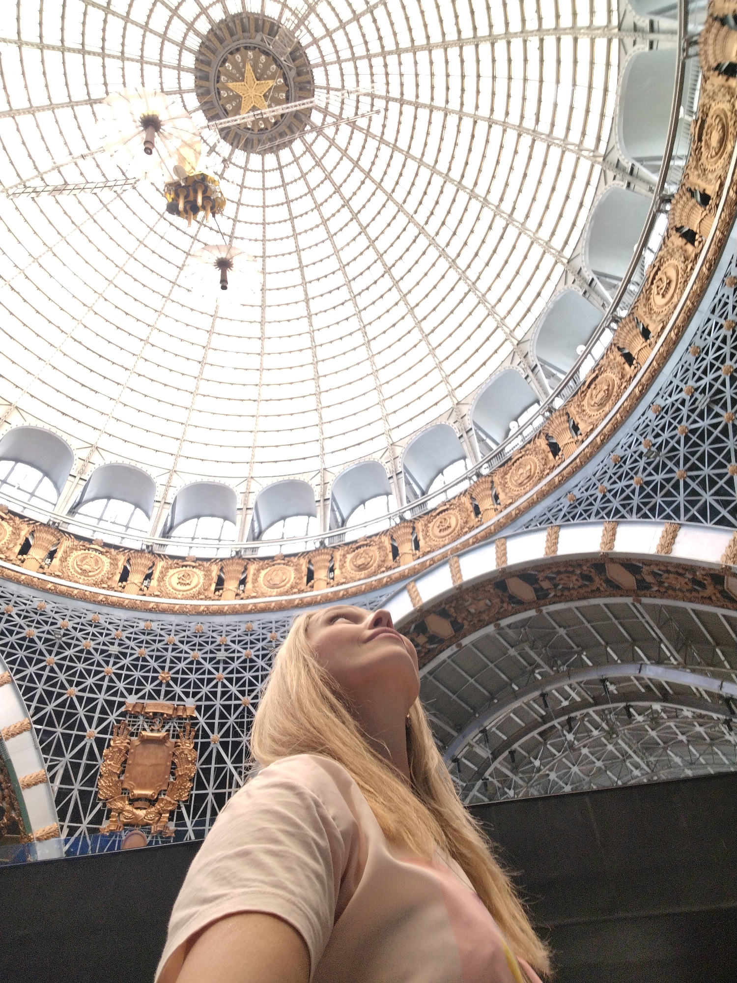 The image depicts a woman looking up at the dome of the Cosmonautics Museum in Moscow, Russia. The woman, with blonde hair and wearing a light pink t-shirt, is positioned in the foreground, partially obscuring the lower part of the frame. The dome, made of glass and metal framework, is intricately designed with a star emblem at its apex and decorative golden elements along its circumference. Several hanging light fixtures are visible within the dome. The architecture suggests a grand and possibly historical interior space.