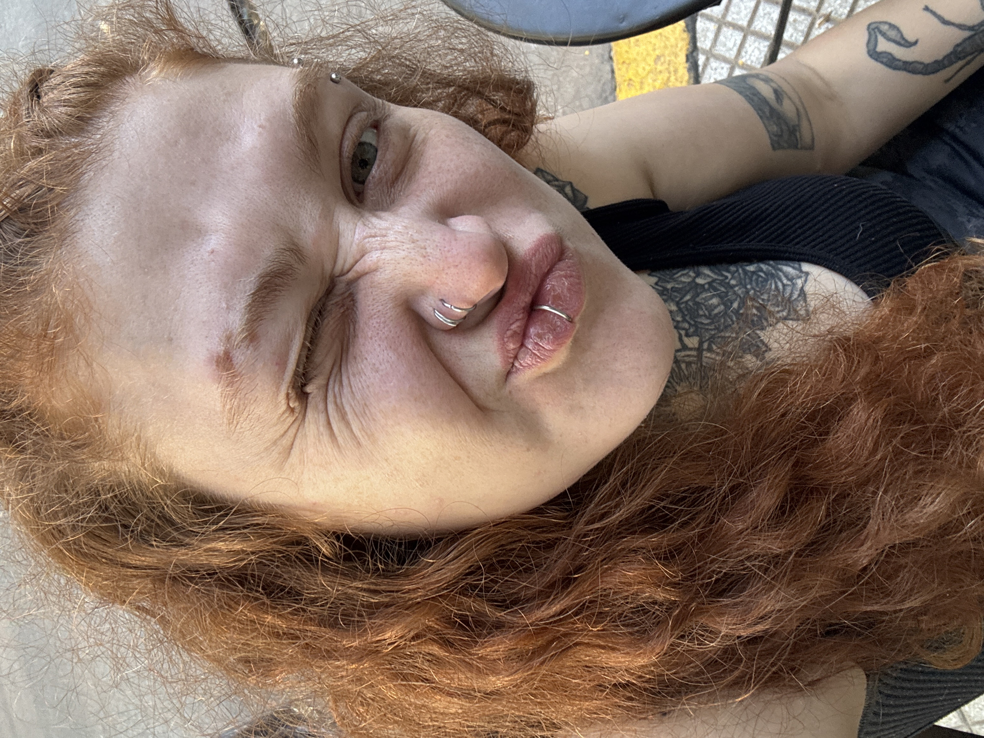 The image is a close-up, top-down portrait of a person with fair skin and long, wavy, reddish-brown hair. The subject is making a playful, expressive face, winking strongly with their right eye, scrunching their nose, and pursing their lips into a pout. Their eye color appears to be green or hazel, and subtle freckles are visible across their nose and cheeks.
The person has multiple silver-colored body piercings: a barbell in their left eyebrow, two hoops in their nose, and a single hoop in their lower lip. Their lips appear somewhat dry or chapped.
They are wearing a black, ribbed top or tank top. Visible tattoos include a stylized, abstract design on their left arm, and a detailed floral or rose-patterned tattoo on their left shoulder/chest area.
The bright, natural lighting suggests the photograph was taken during the daytime in an outdoor setting. A blurred background shows hints of a textured surface, possibly concrete or pavement, with a yellow line, suggesting a street or public space. The overall mood is casual and expressive. Although the provided context is Buenos Aires, Argentina, the image itself does not contain explicit visual cues to confirm this location. There is no visible text in the media file.