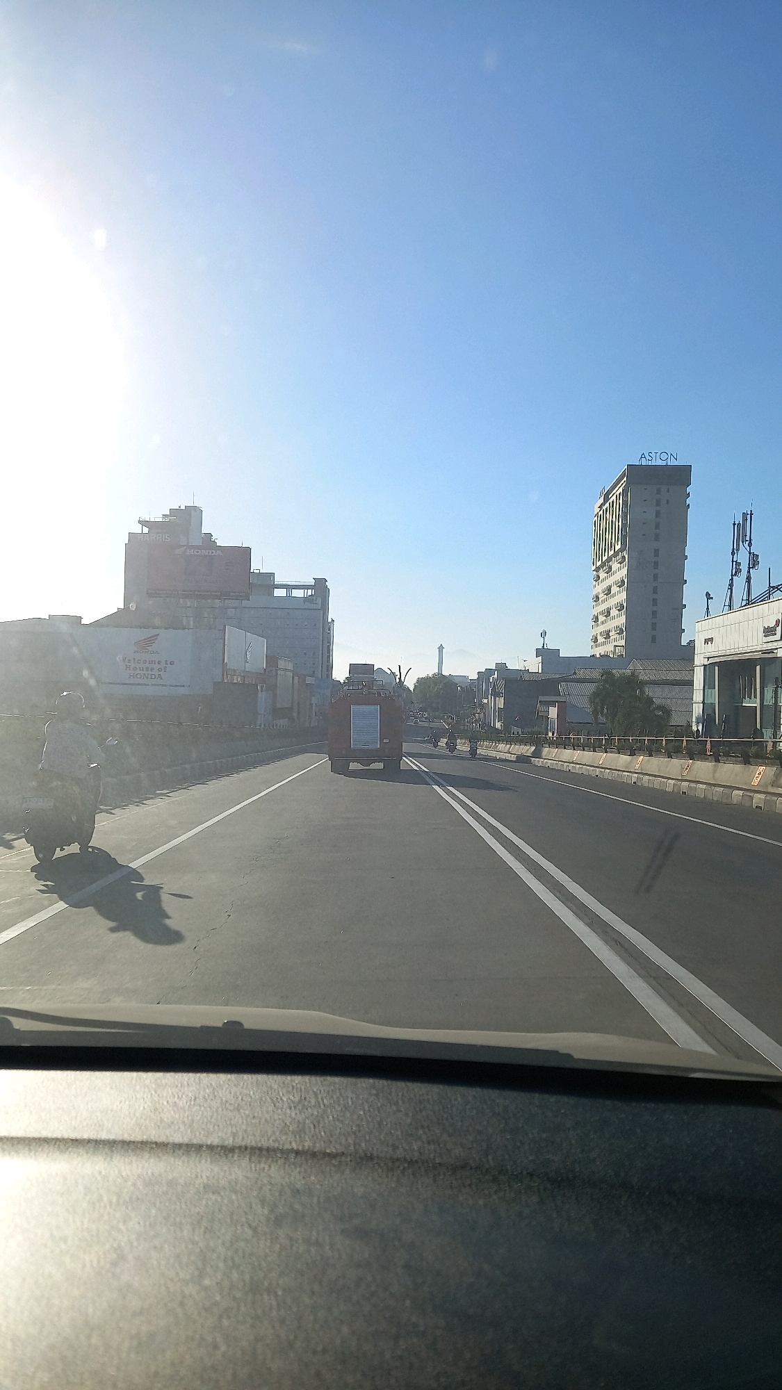 An eye-level, wide shot captures a street scene in Surakarta, Indonesia, on a clear, sunny day. The sun is bright and high in the sky, casting long shadows across the road and the surrounding environment. The perspective is from a vehicle looking forward, with the dashboard visible at the bottom of the frame.
In the foreground, a motorcycle with a rider is moving away from the camera on the left side of the road. The rider is wearing a helmet and a patterned shirt, and the motorcycle is dark in color.
The road itself is asphalt with white lane markings. In the middle of the road, a red vehicle, possibly a fire truck or a utility vehicle, is traveling in the same direction as the camera. It has a tall structure on its back with some poles or pipes.
To the right of the road, there is a concrete barrier. Beyond the barrier, various buildings are visible. The most prominent is a tall, modern building with the word "ASTON" written in blue letters at the top. To the left, a multi-story building displays a large red advertisement with the "HONDA" logo and text reading "Welcome to House of HONDA." Other buildings and structures, including antennas and a minaret-like tower in the distance, suggest an urban environment. The sky is a clear, bright blue. The overall impression is of a busy street in a city under bright sunlight.