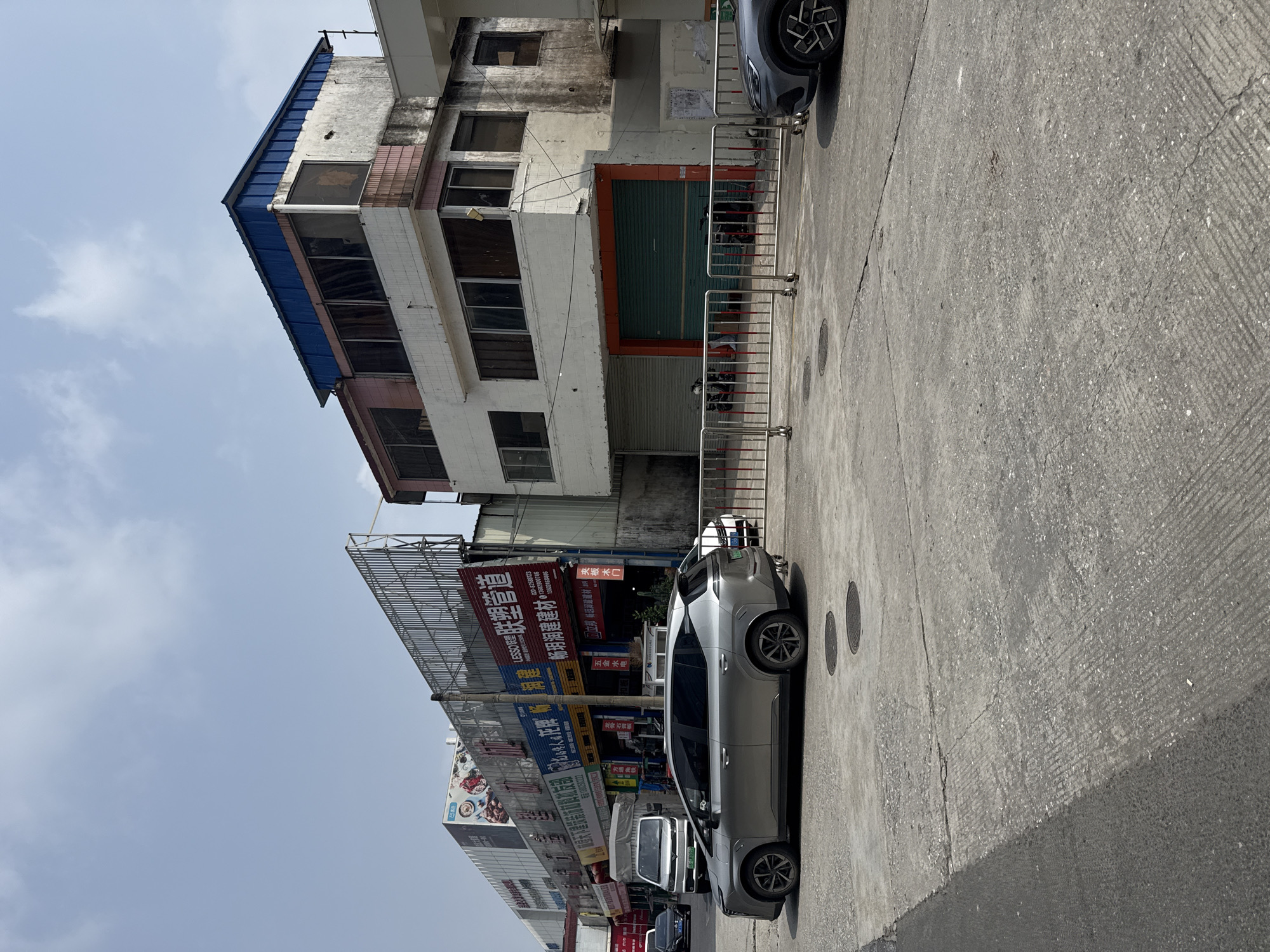 In Tianhe District, China, a street scene unfolds under a bright, partly cloudy sky. A row of commercial buildings lines the right side of the street, with some appearing older and weathered, showing peeling paint and discolored facades. One building has a prominent blue-tiled roof. The ground floor of some buildings features large, metal roll-up doors, one of which is painted a distinctive teal green and outlined with orange. In front of these buildings, a metal barrier separates the sidewalk from the street.
The street itself is paved with concrete, showing cracks and wear. Several vehicles are present: a silver SUV is parked in the foreground, facing left. Further down the street, a light-colored van is parked, and to its right, a dark grey car is partially visible. Traffic lights and street signs are also visible on poles. On the left side of the image, a long, multi-story commercial building stretches into the distance, adorned with large advertisements and signage, some of which are in Chinese. The overall impression is of a functional, busy commercial area.