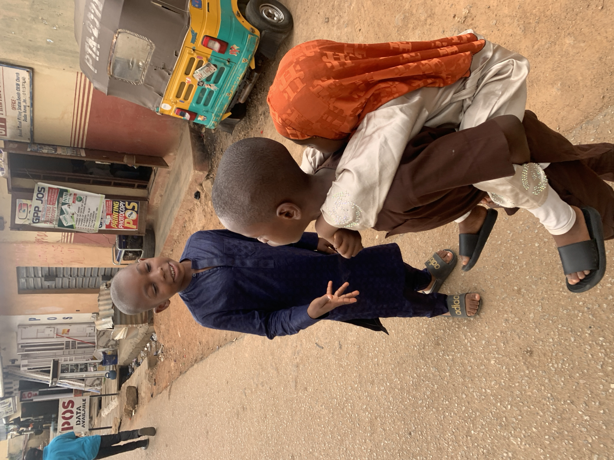 The image shows three children in what appears to be a street scene in Jos, Nigeria. One child in a navy blue tunic is standing, smiling and making a hand gesture. Two other children are in closer proximity to each other, one wearing an orange head covering and the other in brown and white clothing. Next to the children is a yellow and turquoise "keke", a type of motorized tricycle taxi. The backdrop includes building fronts with signs indicating services like "Printing Photocopy", "POS", and "GPP JOS". Another sign references "Band Tatal Filling Station Beside COCIM Church, Dadin-Kova, Jos", which identifies the location in Jos. A man in a blue shirt and black pants is standing in front of the POS shop.