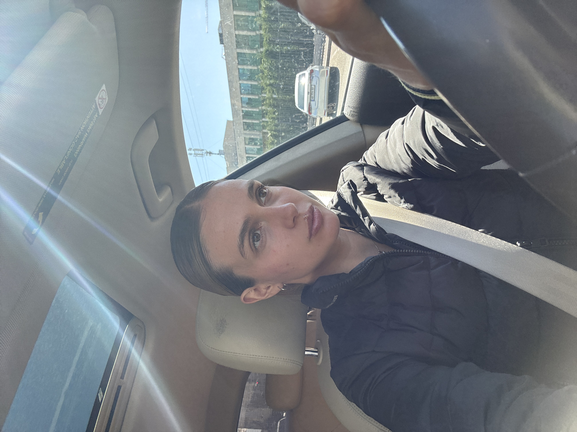 A medium shot depicts a person inside a car. The person, likely female, has light skin, brown hair pulled back, and is wearing a black quilted jacket and a seatbelt. The car interior is beige. The person is looking toward the camera. Outside the car window, buildings and a silver-colored vehicle can be seen. The location appears to be urban with a high-rise building in the background. The city and country are Unknown, Unknown.