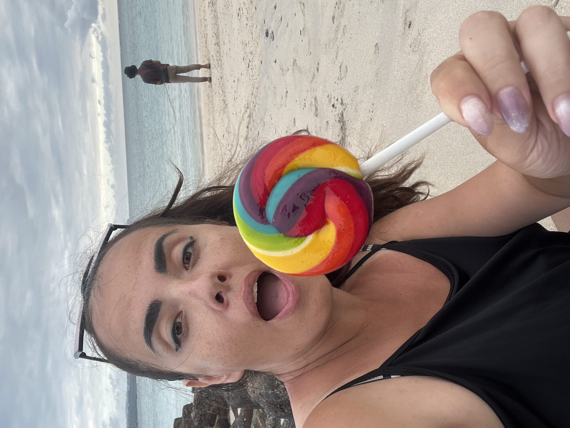 A woman is taking a selfie on a beach in Kuta, Indonesia. She is holding a colorful lollipop up to her mouth. She is wearing a black tank top. In the background, the ocean is visible, and a person is standing on the beach facing away from the camera. The sky is cloudy.